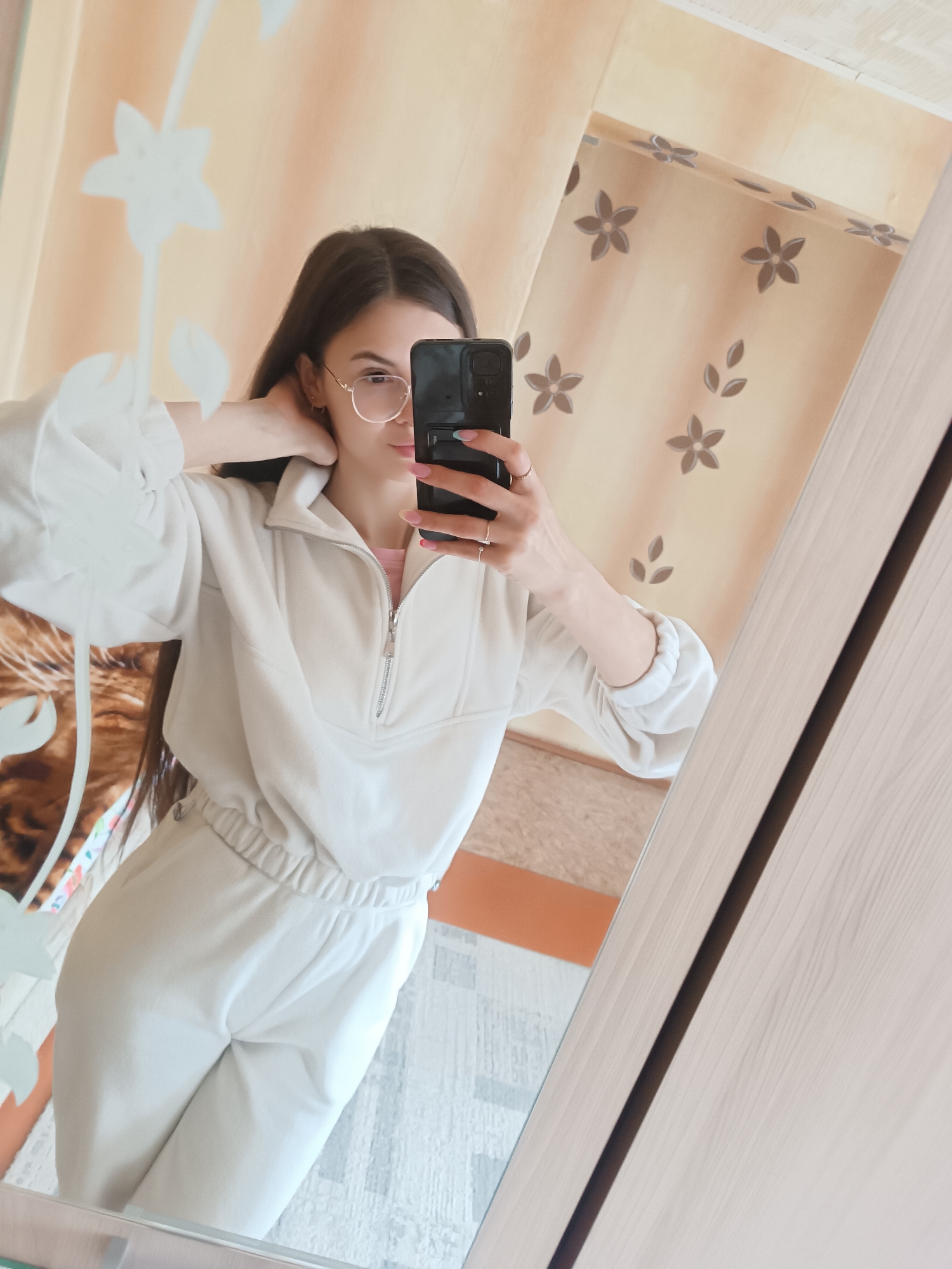 A young woman is captured in a mirror selfie, reflecting her image in what appears to be an indoor setting, likely a bedroom or living room. She has long dark hair and is wearing glasses with thin, light-colored frames. Her gaze is directed towards her smartphone, which she holds in her right hand, poised to take the picture. Her visible left hand is positioned behind her neck or touching her hair.
She is dressed in a casual, light cream or off-white two-piece loungewear set, consisting of a sweatshirt with a half-zip neckline and matching pants with an elasticized waist. Her fingernails have light pink polish, and she wears several rings on her fingers. The black smartphone in her hand features a visible camera module with multiple lenses.
The background reflected in the mirror shows light-colored wallpaper adorned with a repeating pattern of dark brown/beige stylized flowers and leaves. The floor appears to be a light-toned wooden or laminate surface, with a different section of flooring or a rug visible. On the far left, a blurred, textured object, possibly a furry blanket or throw, is partially visible. A distinctive white decorative floral pattern is present on the mirror's surface or an object positioned in front of it. The mirror itself has a light wooden frame on its right side. The lighting suggests it is daytime, with soft ambient light. There is no visible text in the image.