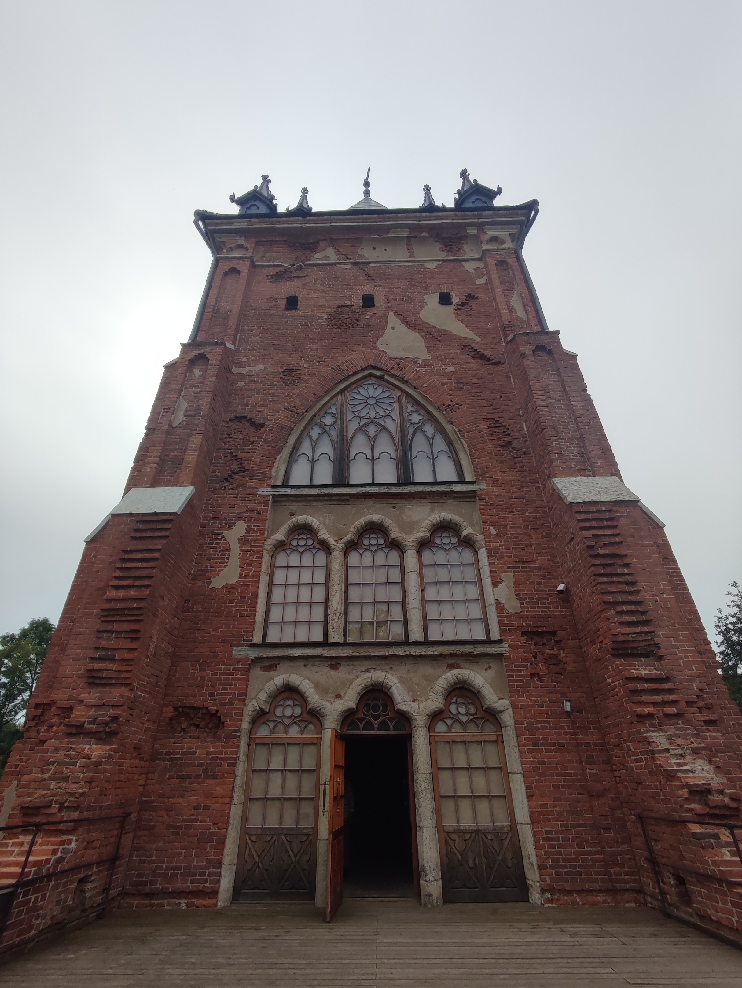 An exterior, low-angle shot captures the grand facade of the Chinese Palace's kitchen tower, a prominent landmark in Catherine Park, Pushkin, Russia. The imposing brick structure stands tall against an overcast sky, its weathered facade telling a story of time. The tower's architecture features a series of arched windows, each intricately decorated, with a particularly large, rose-like window gracing the upper section. Below this, three smaller, arched windows are arranged in a row. The entrance to the tower is marked by a pair of wooden doors, one of which stands ajar, hinting at the secrets within. The brickwork, a rich, earthy red, shows signs of wear and tear, with patches of plaster missing, revealing the underlying structure. The surrounding environment is hinted at by the presence of trees on the left and right edges of the frame, suggesting a park setting. A wooden pathway leads up to the entrance, with a railing on the left side for support. The overall impression is one of historical grandeur and architectural beauty, despite the visible signs of age.