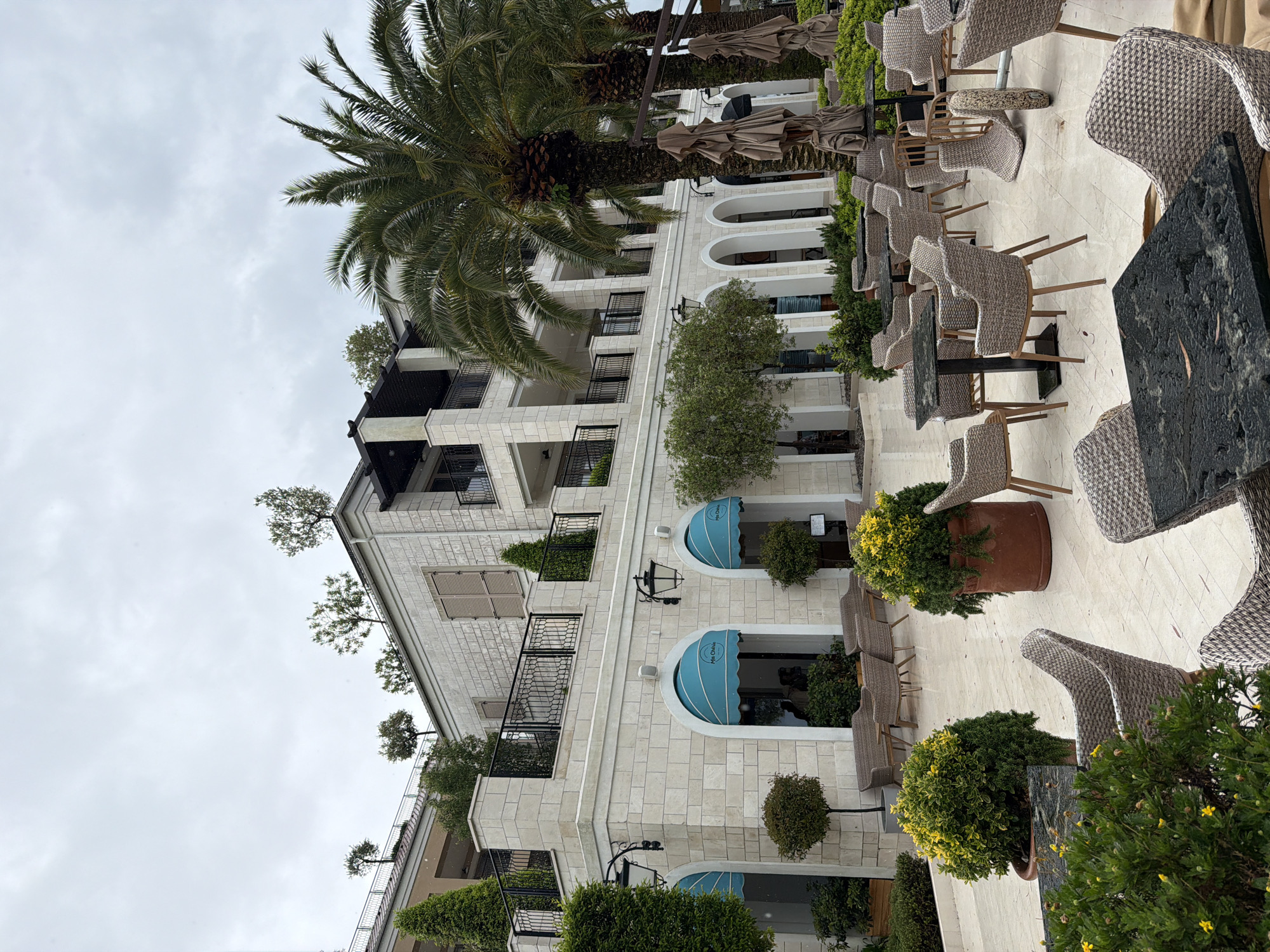 This image shows the outdoor seating area of a restaurant or cafe in Tivat, Montenegro. The area features wicker chairs and tables arranged on a light-colored paved surface. There are several potted plants with green and yellow foliage interspersed among the tables, and an umbrella stands folded in the background. A multi-story building is seen with white stone cladding, arched windows, and wrought-iron balconies. Palm trees are visible, and the sky is overcast.