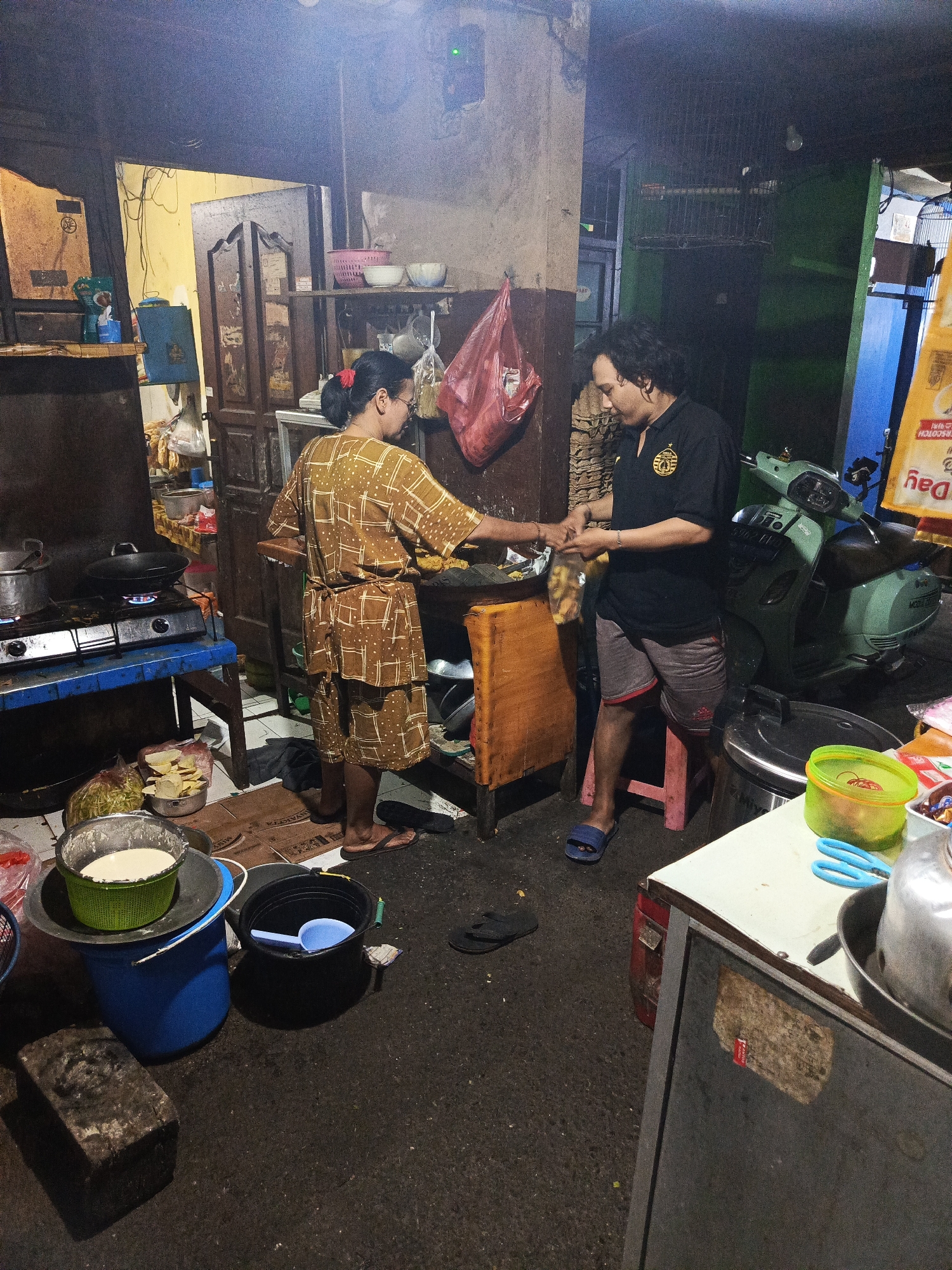 A medium shot captures an indoor scene, possibly a food stall or a residential kitchen, in Central Jakarta, Indonesia. A woman in a brown patterned outfit stands to the left, facing away from the camera, and appears to be preparing food on a gas stove. A man in a black polo shirt and gray shorts stands to the right, interacting with the woman. They are exchanging something, possibly ingredients or money. The environment is cluttered with various items, including cooking utensils, a scooter parked in the background, and what appear to be sacks of goods. The lighting is dim, suggesting it may be evening or the area is not well-lit.