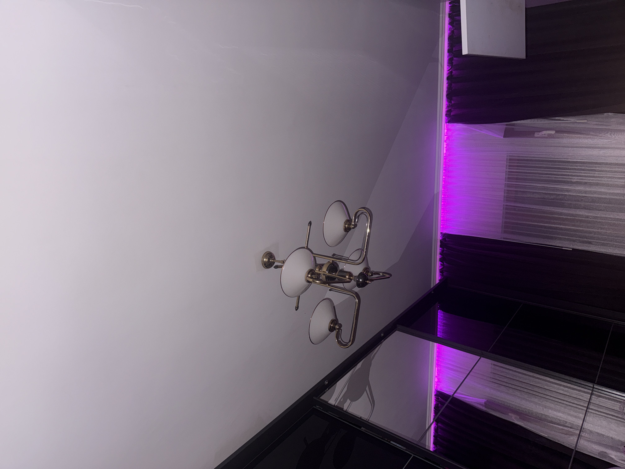 This vertically oriented image captures an indoor setting, likely a modern room or corridor. A prominent brass-toned chandelier, featuring at least five white, bowl-shaped shades, is mounted on a plain, light-colored wall, appearing almost off-white or light grey. Below the chandelier, a highly reflective, dark surface, possibly a glossy floor or a large mirrored panel, reflects the light fixture's silhouette and other elements of the room.
To the right side of the frame, a vibrant strip of purple LED lighting casts a strong glow. This light illuminates what appears to be a window or a recessed alcove, fitted with blinds or curtains that are partially covered by translucent protective plastic sheeting, suggesting recent installation or renovation. Above this illuminated section, a dark, possibly paneled wall extends upwards, with a rectangular white panel or fixture attached to it.
No people are present, and no specific activity is taking place. The artificial lighting, particularly the striking purple strip, indicates an indoor environment where the natural time of day is not discernible. The combination of the chandelier and modern LED strip suggests a contemporary design aesthetic. There is no visible text in the image. This scene is located in Stara Zagora, Bulgaria.