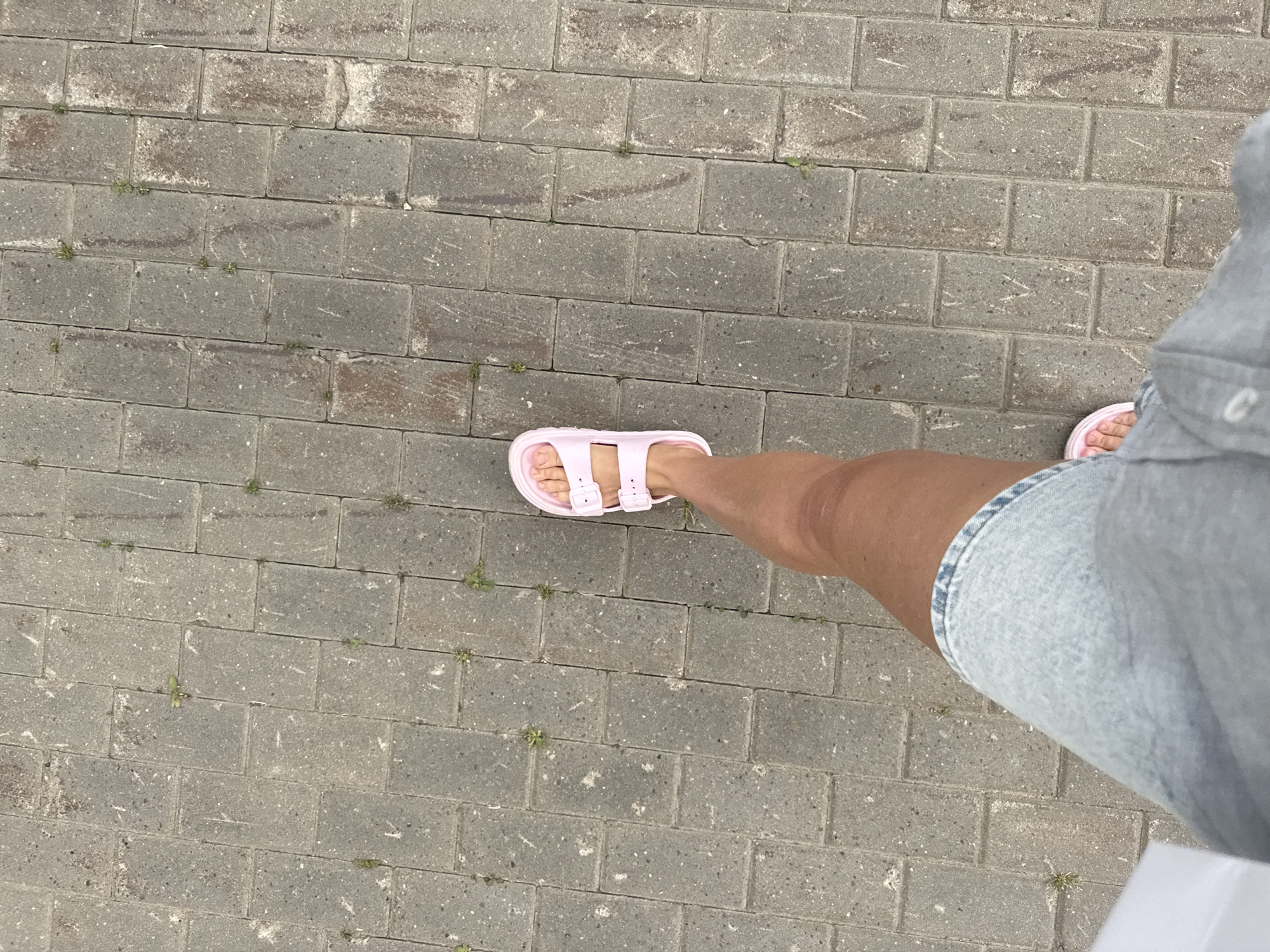 The image shows a person's legs and feet walking on a paved walkway. The walkway is made of rectangular gray bricks laid in a running bond pattern. Small patches of green grass are growing in the gaps between the bricks. The person is wearing light blue denim shorts and pink slip-on sandals. The perspective is from above, looking down at the person's legs and feet. The location is the city of Ankudinovka, Russia.