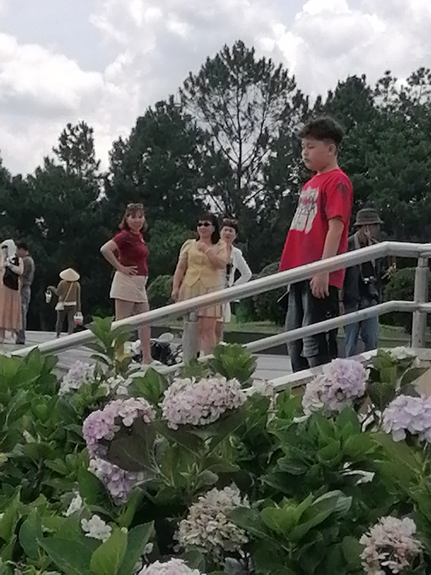 The image depicts a group of people on a paved walkway, with lush green foliage and hydrangeas in the foreground. The walkway is bordered by a metal railing. In the background, tall trees form a dense canopy. Several individuals are visible: a young boy in a red t-shirt stands prominently in the right side of the frame, looking down. To his left, a woman in a yellow dress and sunglasses is partially visible. Further left, a woman in a red top and white skirt stands with her hand on her hip, looking towards the camera. Behind her, another woman in a yellow outfit and sunglasses is visible. To the far left, a woman in a beige dress and dark vest is seen from the side. A person wearing a conical hat and a brown jacket is visible in the mid-ground, carrying a bag. The overall atmosphere suggests an outdoor gathering or a tourist location. This scene is likely at the Thien An Monastery in Da Lat, Vietnam, known for its beautiful gardens and scenic views, often frequented by tourists.