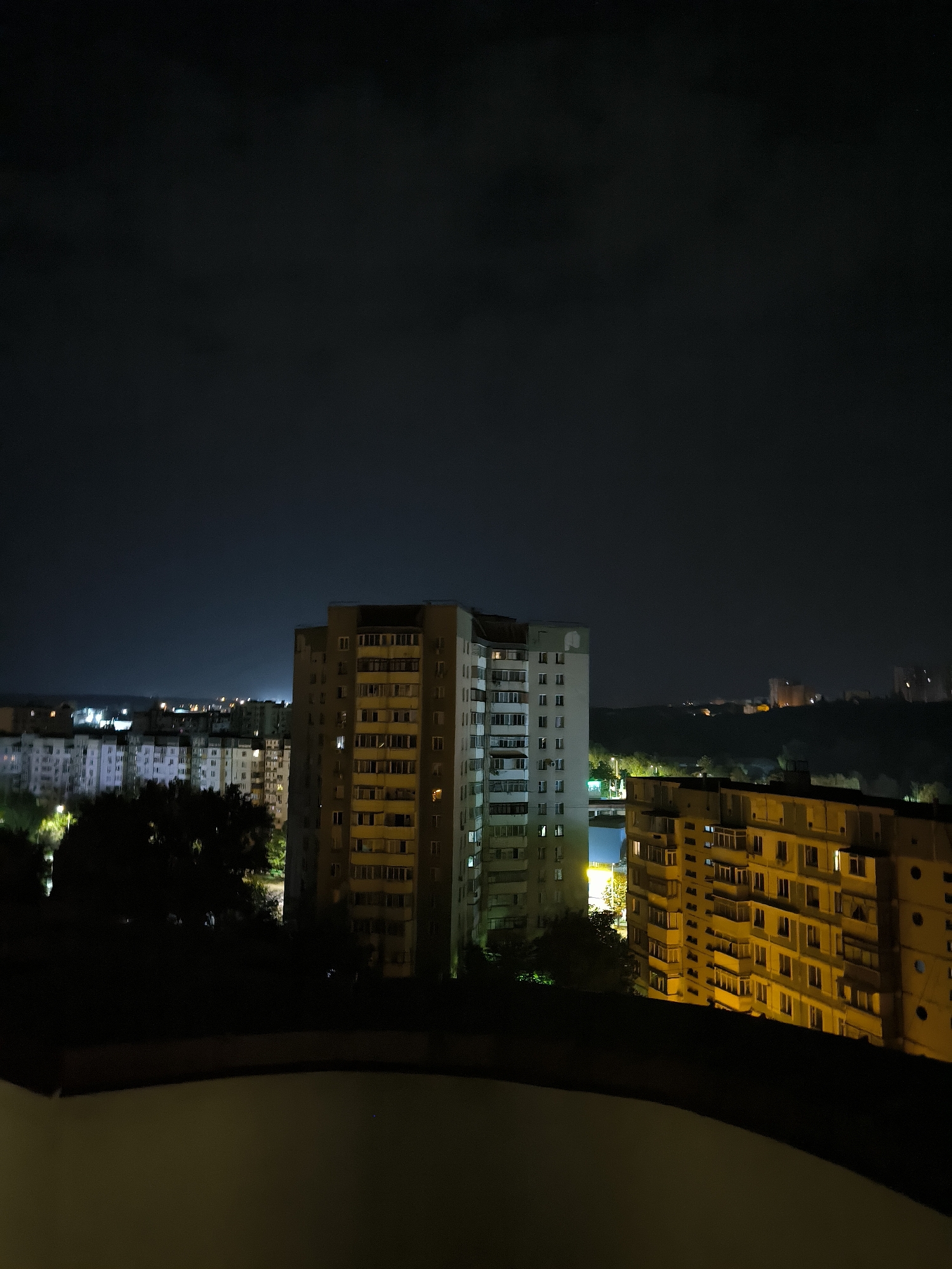 This image captures a nighttime view of a residential area in Chișinău, Moldova. The foreground features a dark balcony railing, obscuring the bottom portion of the scene. Beyond this, several multi-story apartment buildings are visible, illuminated by internal lights and streetlights.
The buildings are characteristic of Soviet-era architecture, with repetitive window patterns and balconies. In the mid-ground, a prominent tall building with a lighter facade stands out, its windows glowing with a warm yellow light. To its right, another apartment block, primarily yellow, shows a similar pattern of illuminated windows. Further in the background, more buildings recede into the darkness, with scattered lights suggesting a sprawling urban landscape.
The sky is dark and cloudy, with faint hints of lighter patches indicating city lights reflecting off the clouds. Trees are visible in the mid-ground, silhouetted against the illuminated buildings. The overall impression is of a typical urban scene at night, with the lights of the city providing the main source of illumination against the dark sky. There are no people or vehicles clearly discernible in the image.