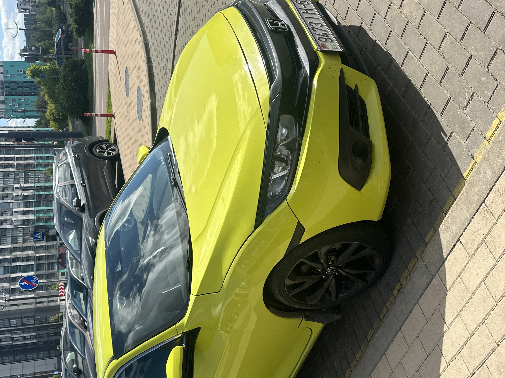 A slightly rotated close-up shot shows a neon yellow Honda Civic parked on a brick-paved curb in Бараўлянскі сельскі Савет, Belarus. The car is positioned partially on the sidewalk, casting a shadow on the light-colored bricks. The car's front passenger side is prominently featured, including the front wheel with a black rim and the Honda logo. The windshield reflects the sky and surrounding buildings, including apartments. In the background, other parked cars are visible, and a building with multiple floors is visible. Red and white safety bollards are visible. A road sign is also visible on the side of the road.