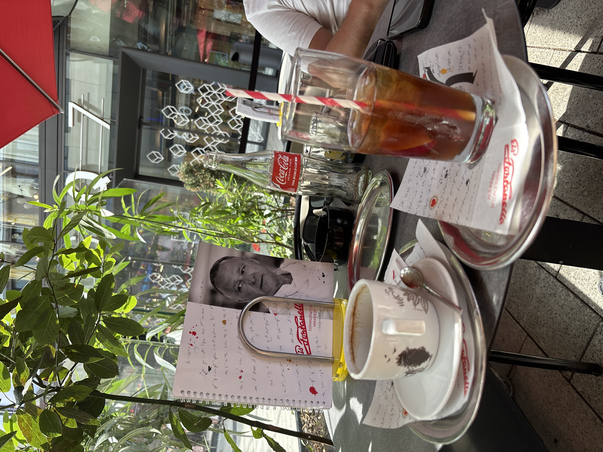 The image shows a table at an outdoor cafe with a variety of items. In the foreground, there is a white coffee cup with a saucer and a small spoon. Next to it, there is a notebook with a black and white photo of a man on the cover, and handwritten text. A Coca-Cola bottle and a glass with a red and white striped straw are also visible on the table. The scene appears to be set during the daytime, with sunlight casting shadows. The overall atmosphere suggests a relaxed cafe setting, possibly in a city given the architectural elements visible in the background. The text "Coca-Cola ORIGINAL TASTE FIFA WORLD CUP" on the bottle indicates a connection to a past sporting event. The text on the napkin and notebook mention "Fontanella EISMANUFAKTUR MANNHEIM," suggesting the cafe is likely located in Mannheim, Germany, and specializes in ice cream.