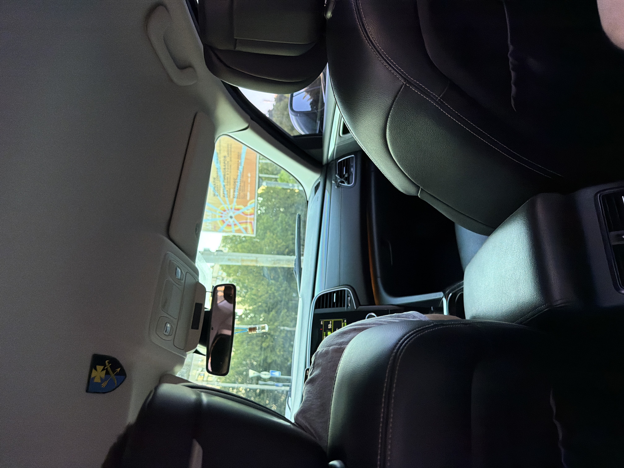 The image captures the interior of a car, looking forward from the back seats. A dark-colored leather headrest is prominently in the foreground. To the left, another dark-colored leather headrest and the shoulder of a person wearing a grey t-shirt are visible. Above, a shield emblem with yellow and blue elements, featuring a cross and what appears to be a sword or spear, is attached to the car's ceiling. A rearview mirror hangs from the ceiling, reflecting the interior and a glimpse of the outside. A small glass vial on a string is suspended from the mirror. The car's dashboard is visible, with a central screen displaying some information and a vent below it. A portion of the windshield shows an outdoor scene with trees and a billboard with Ukrainian text. The overall setting suggests a daytime drive within a city.