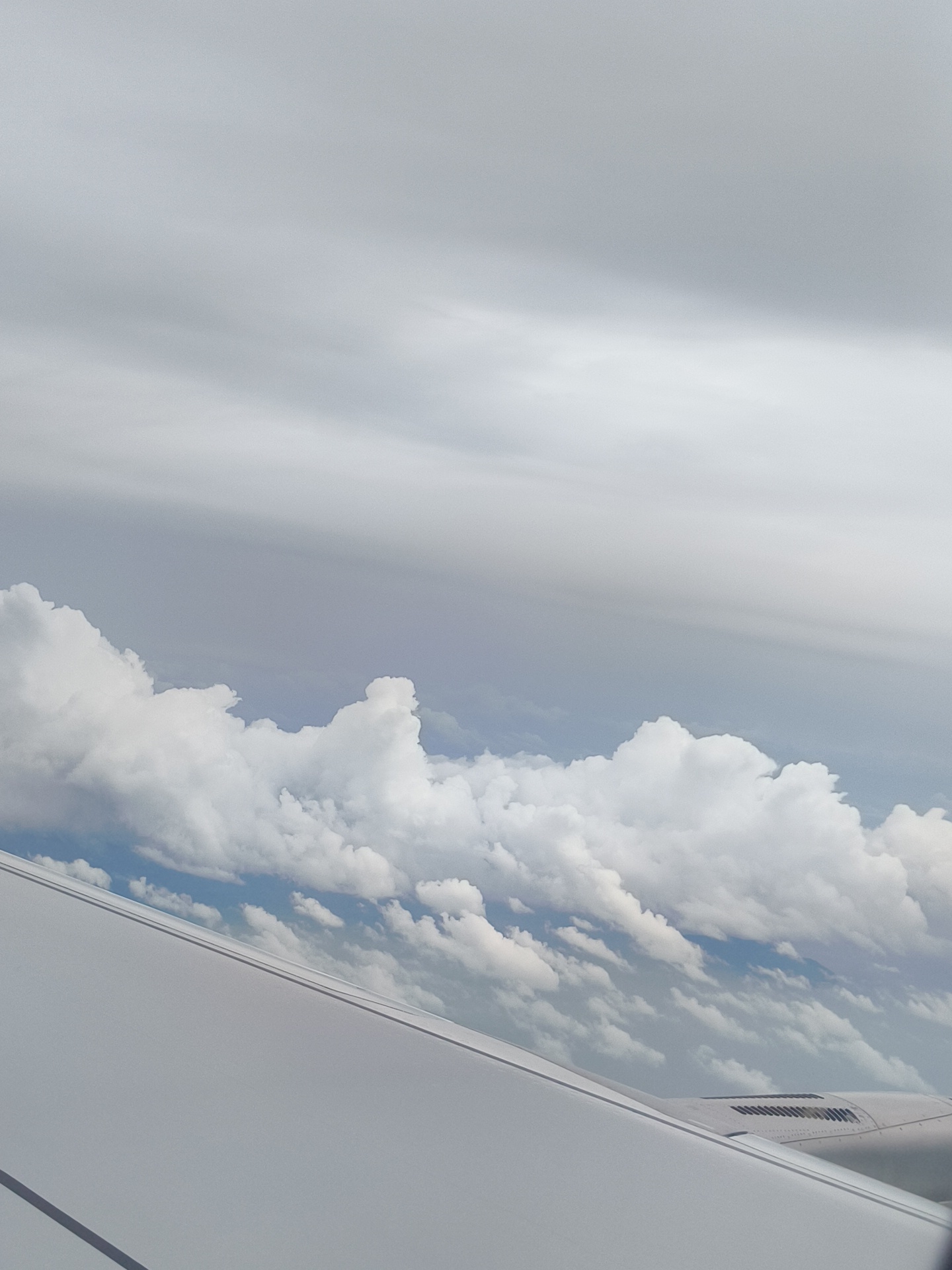 An aerial view from an airplane window shows a vast expanse of clouds under a pale sky. The clouds are white and fluffy in the foreground, with some appearing more wispy and distant in the background. Below the clouds, patches of blue sky are visible. The lower portion of the image is dominated by the white surface of an airplane wing, with subtle lines indicating structural details. No people or specific landmarks are visible. The scene depicts a typical flight environment.