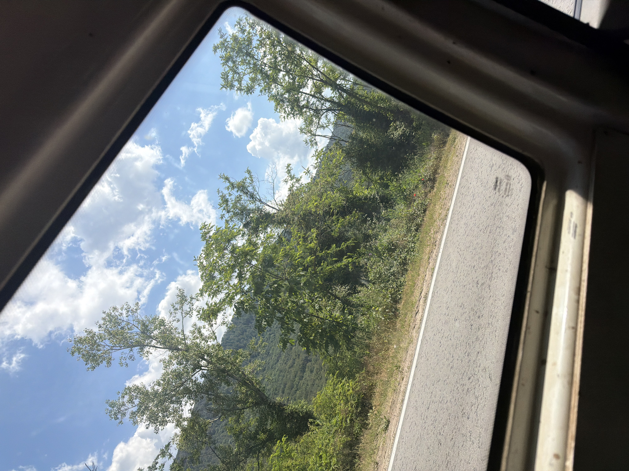 A view from inside a vehicle looking out a window at a tree-lined roadside and a forested hillside under a bright, cloudy sky. The road, paved with asphalt and marked with a white line, runs horizontally across the lower portion of the image. Lush green foliage, consisting of trees and bushes, borders the road and climbs the gentle slope of the hill in the background. The sky is a vibrant blue, dotted with scattered white cumulus clouds. The interior of the vehicle is visible in the foreground, framing the outdoor scene. There are no people visible in the image. This scene appears to be located on the outskirts of Balgarene, Bulgaria, along a rural road surrounded by natural greenery.