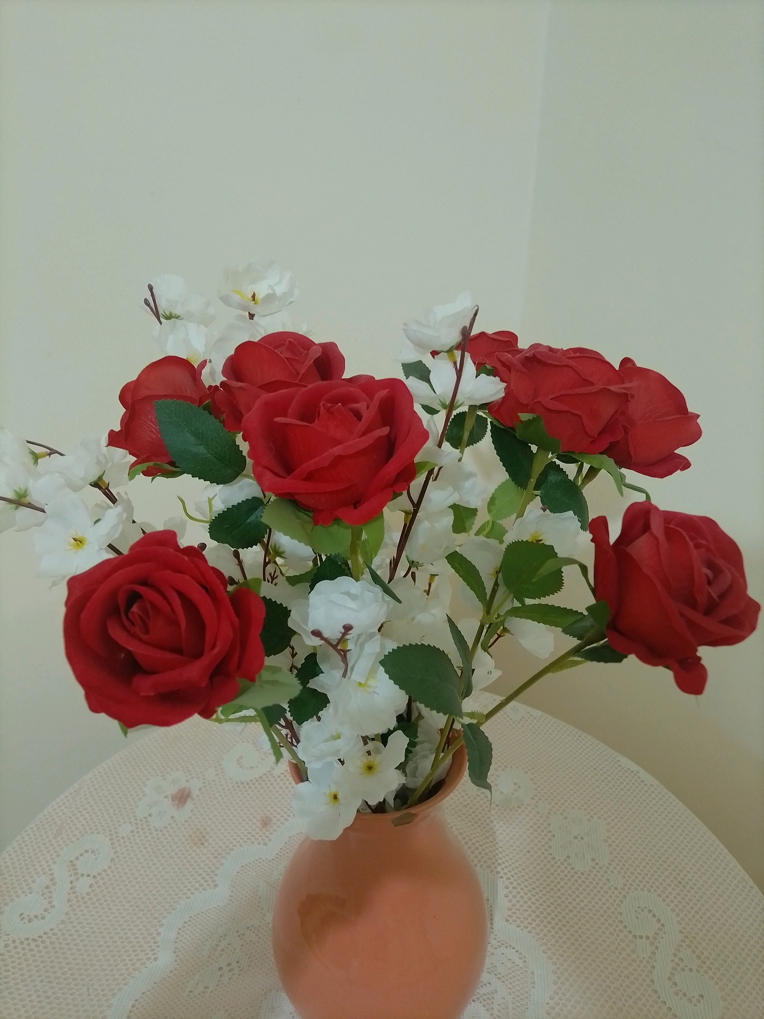 A bouquet of red roses and white flowers is arranged in an orange vase on a lace-covered table. The flowers are artificial and appear to be made of fabric or plastic. The background is a plain, light-colored wall. The scene is indoors, and the lighting suggests it is daytime. There are no people visible in the image. The location is not specified within the city of Amman, Jordan, nor is there any information to suggest this location.