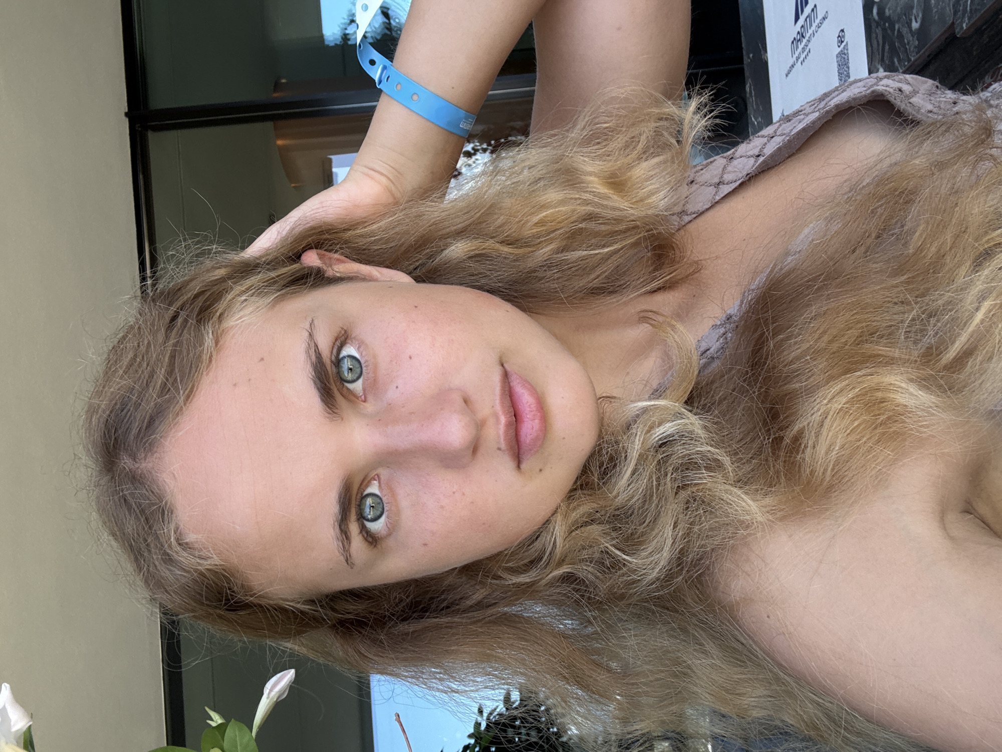 A young woman with blonde, wavy hair and blue eyes is taking a selfie outdoors. She has her hand in her hair, and is wearing a light-colored top. A blue wristband is visible on her right wrist. The background includes some greenery and a building with large windows. The image appears to be taken in the Vlora, Albania area.