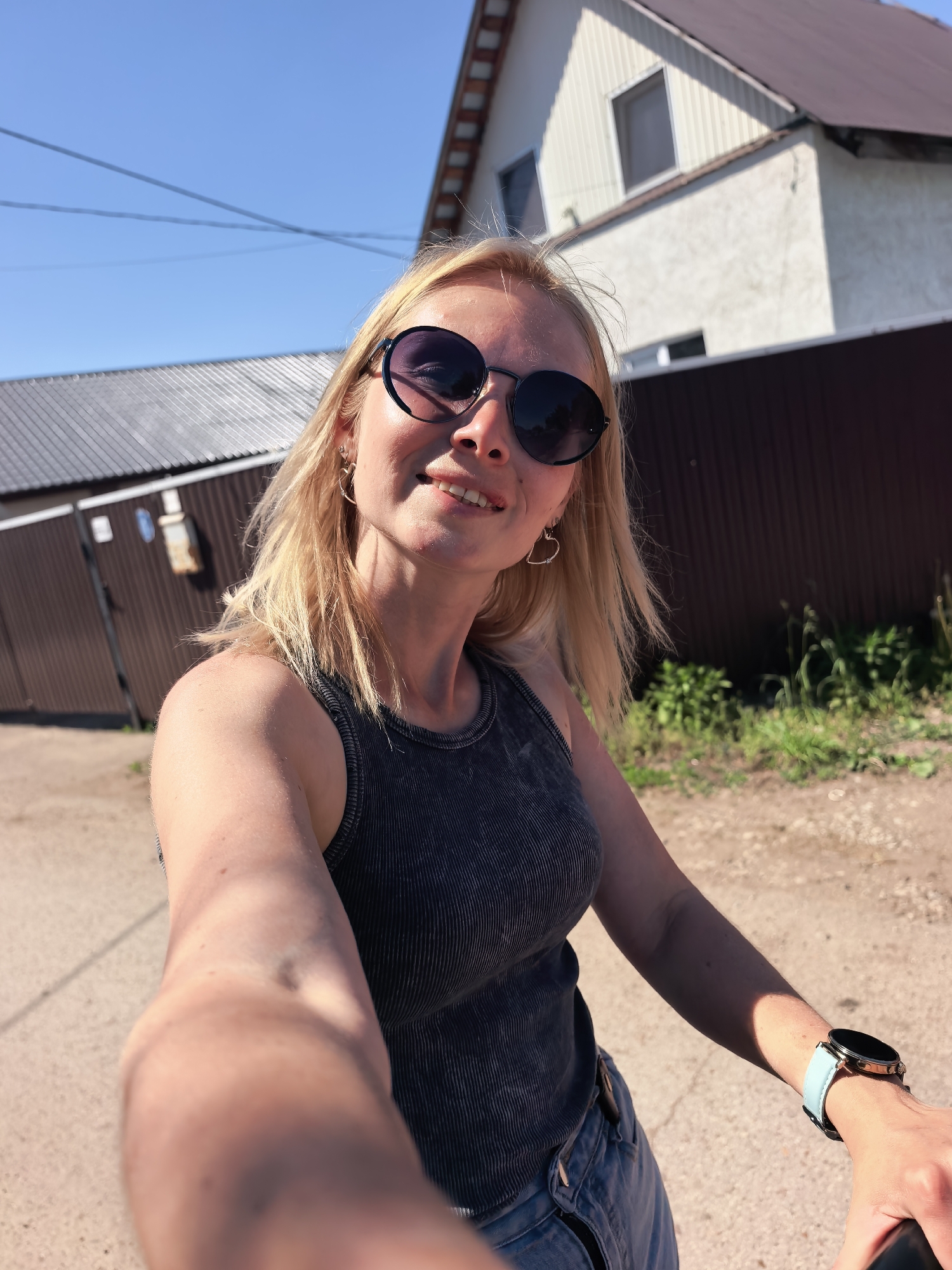 A woman with blonde hair and sunglasses takes a selfie in Sterlitamak, Russia. She wears a dark gray tank top and blue jeans. She is on a street with a brown fence, green grass, and a house in the background. The sky is clear and blue. She is wearing a smartwatch with a light blue band.