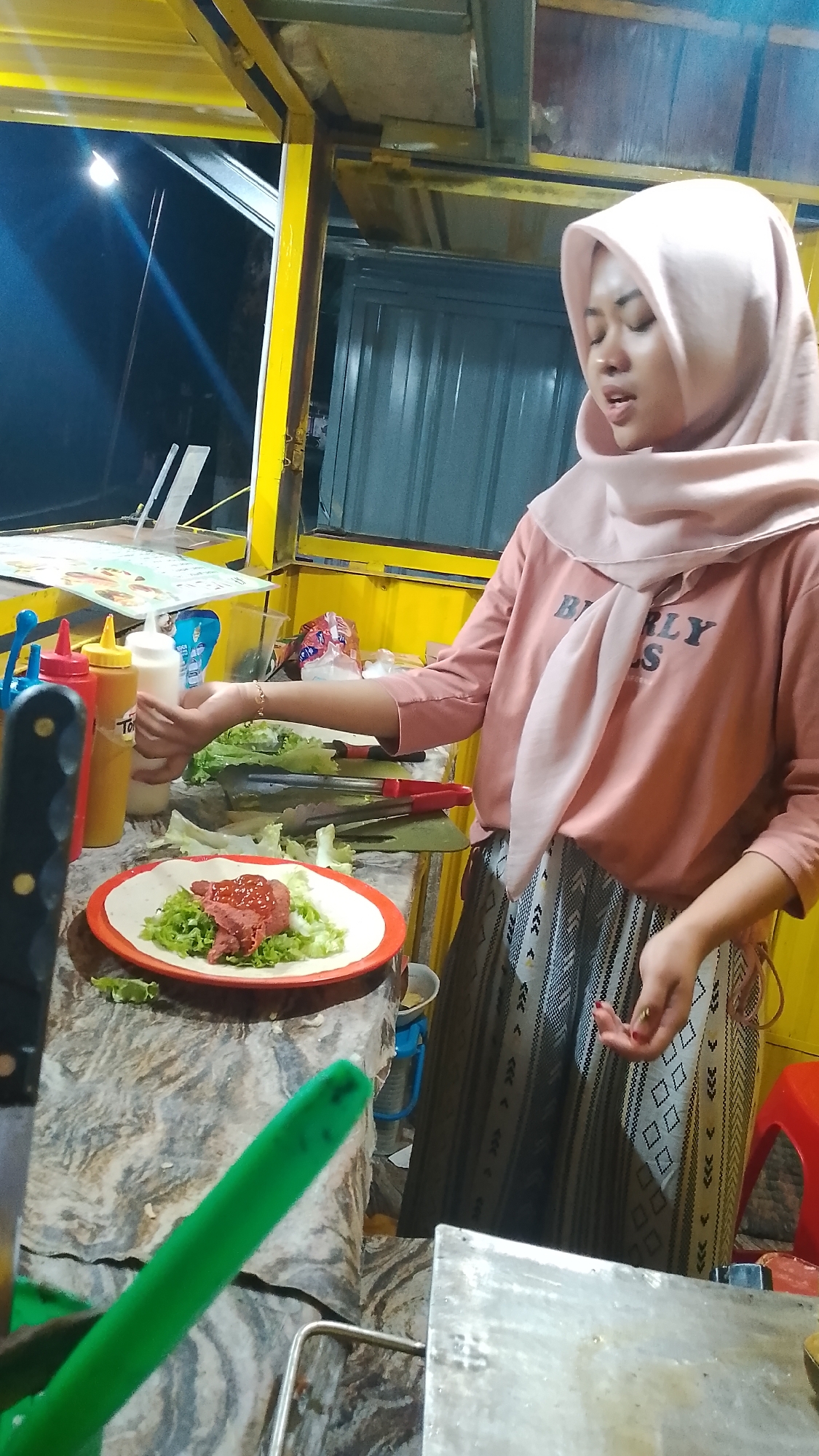 A young woman wearing a light pink hijab and a salmon-colored t-shirt with the words "BEARLY" and "S" printed on it, and a patterned skirt, is preparing food at an outdoor food stall in Karanglangu, Indonesia. She is standing behind a counter with a marble-like pattern, which is cluttered with various items including bottles of condiments (red ketchup, yellow mustard, and white mayonnaise), a plate of food, and cooking utensils. On the plate, there is a flatbread topped with lettuce and meat, which appears to be a wrap or burrito. To the left of the plate, a large knife with a black handle is visible, and to the front, a bright green utensil is also present. The food stall has a yellow metal frame and a corrugated gray wall is visible in the background. The lighting suggests it is evening or nighttime, with a bright light source illuminating the scene from the left.