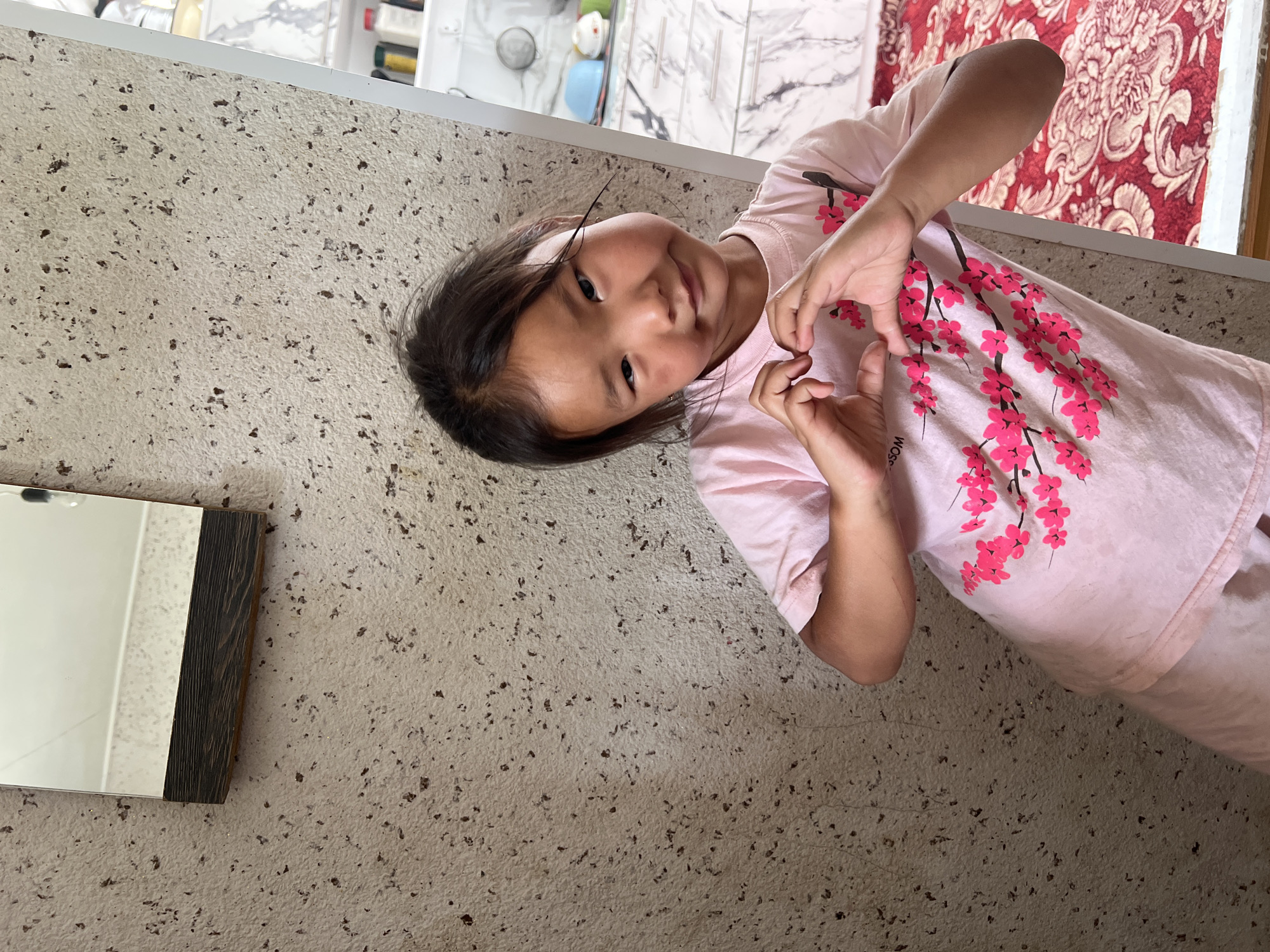 A young girl with dark hair and a light complexion is posing for a photo indoors. She is wearing a pink t-shirt with a pattern of pink cherry blossoms. Her hands are held together in front of her chest, forming a heart shape. The background consists of a textured wall on the left and a cabinet with white drawers and a marble-like pattern on the right. A red carpet with a floral design is visible on the floor. The overall scene is bright and appears to be a casual indoor setting. The location is identified as Kara-Dıykan, Kyrgyzstan.