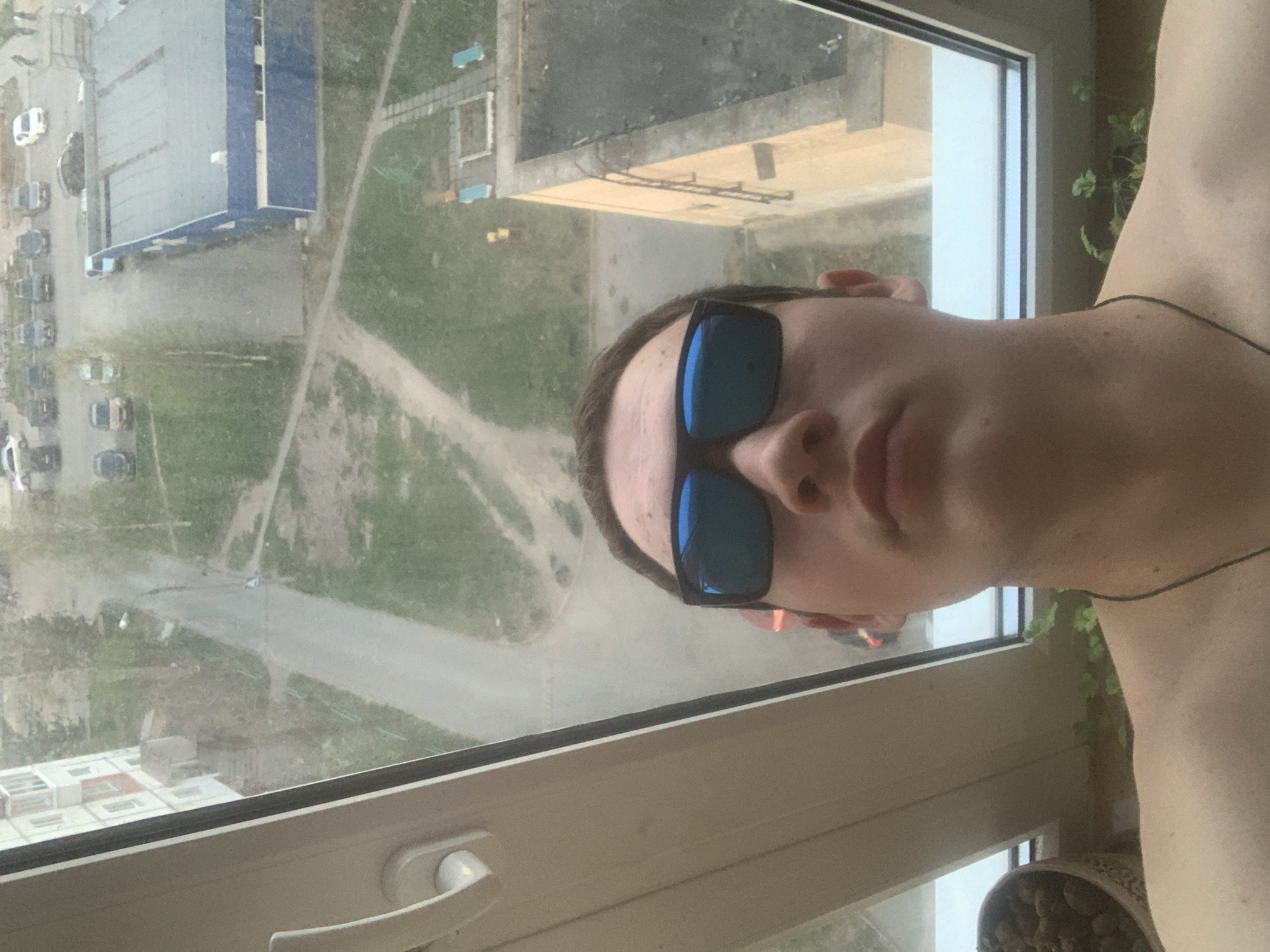 A man wearing sunglasses takes a selfie from inside a building, looking out of a window. Outside the window, the scene reveals a portion of Severodvinsk, Russia. Visible elements include buildings, streets, cars parked along the street, and patches of green space. A window handle and a potted plant are also visible inside the building.