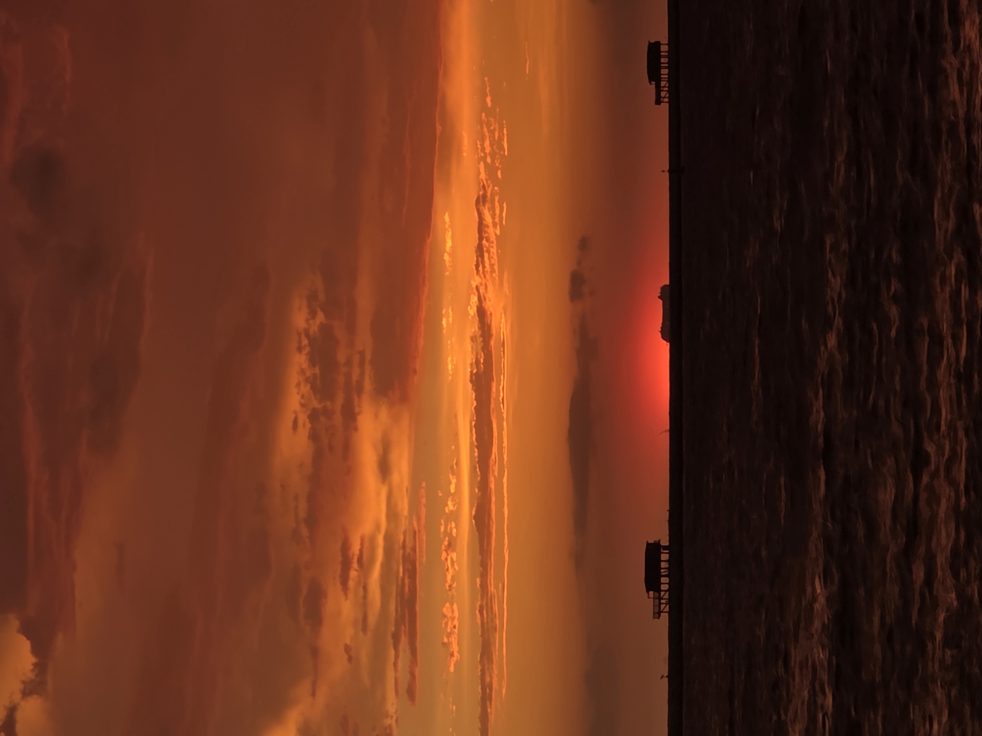 This image depicts a dramatic sunset over the Adriatic Sea, likely viewed from Chioggia, Italy, a coastal city known for its picturesque lagoons and fishing traditions. The sky is ablaze with vibrant hues of orange and red, with a sliver of the sun's disc peeking through the horizon, casting a fiery glow. Wispy clouds, illuminated from below, add texture and depth to the celestial canvas.
In the foreground, the dark, choppy surface of the sea stretches across the frame, reflecting the somber tones of the sky. A thin, dark line on the horizon separates the sea from the sky, and on this line, two silhouetted structures are visible. The structure on the left is a stilted building, possibly a fishing hut or a small dwelling, raised on wooden posts above the water. To its right, a flatter, more elongated silhouette suggests a pier or a breakwater, with a solitary post marking its presence. Further to the right, a second, similar stilted building is also visible, situated on the distant horizon. The overall impression is one of serene isolation and the raw beauty of nature.