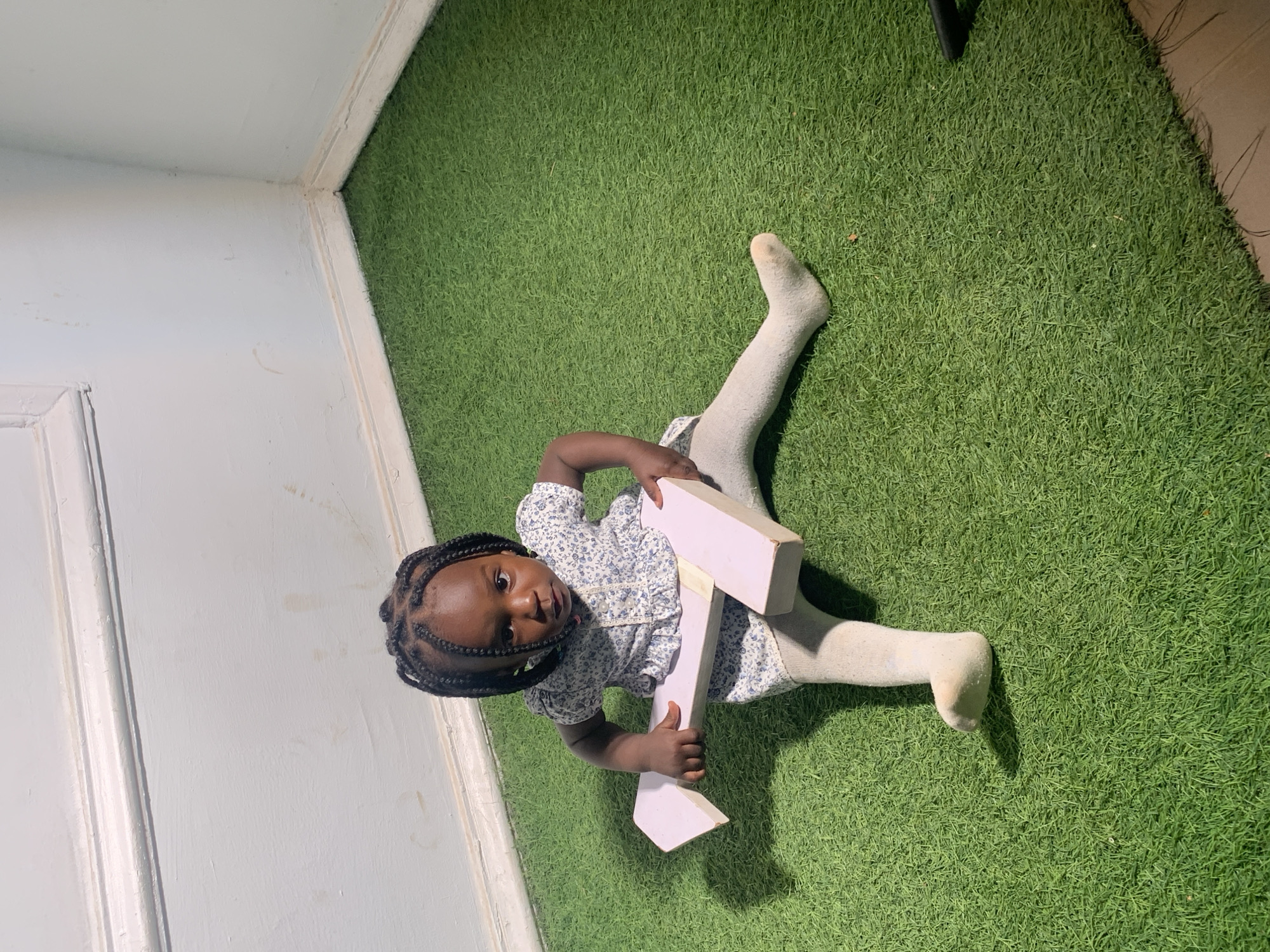 A young Black child is lying on a green, artificial turf surface in what appears to be an interior room. The child is wearing a light-colored, floral-patterned dress and light-colored socks. They are holding a large, light-pink wooden "T" in their hands. The left wall is light blue. The location is Barakin Akawo, Nigeria.