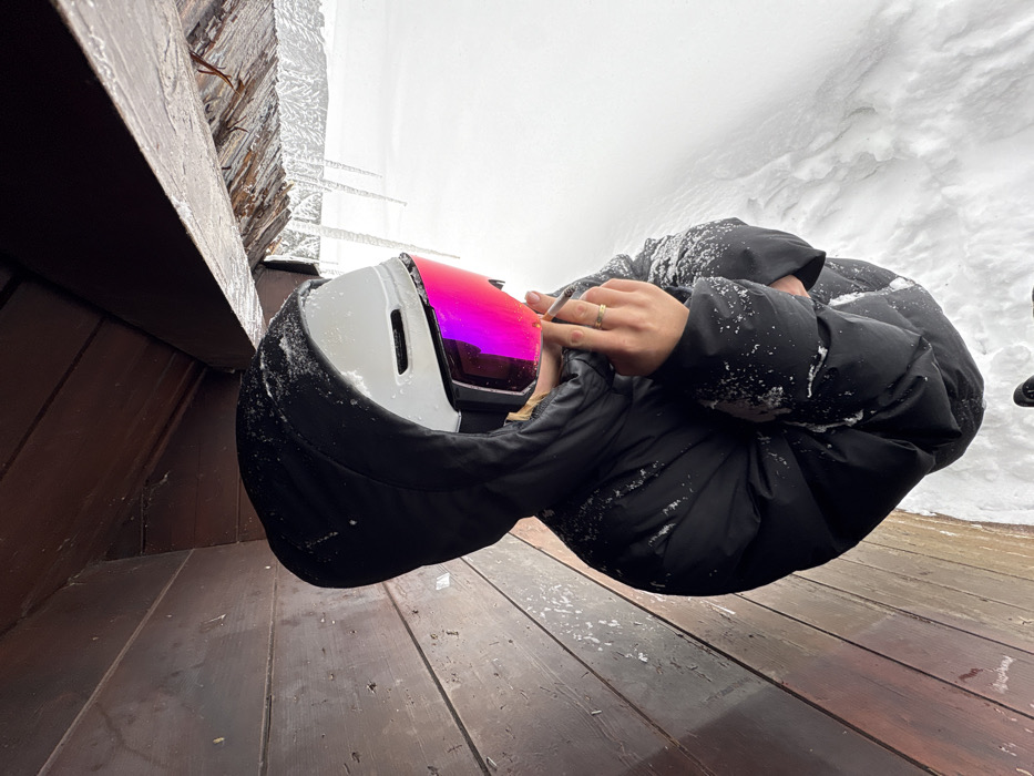 An individual, gender indeterminable, is depicted in an outdoor, snow-covered mountainous environment within the commune of Manigod, France. The person wears a black hooded puffer jacket, dusted with snow, a white ski helmet, and ski goggles featuring reflective pink/purple lenses that obscure the face. Blonde hair is visible beneath the helmet and hood. The individual's right hand is raised, holding a lit cigarette between the thumb and index finger; a plain gold band ring is present on the right ring finger. The person is positioned partially under a dark wooden overhang or shelter, characterized by horizontal wooden planks to the left and an overhead wooden beam with multiple hanging icicles. Beyond the wooden structure to the right and in the background, a vast, undisturbed expanse of white snow is visible.