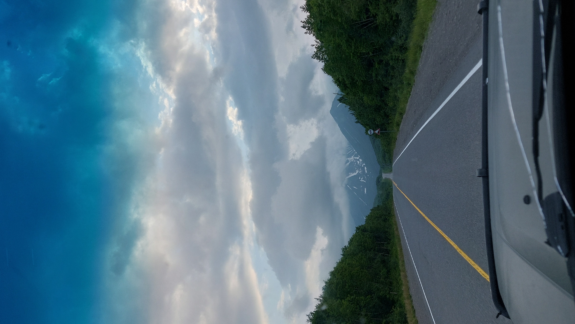 The image shows a view from inside a vehicle looking out at a road leading towards a mountain range under a cloudy sky. The road is asphalt with a yellow double line dividing the lanes and a white line on the right shoulder. Dense green trees line both sides of the road. Snow-capped mountains are visible in the distance, partially obscured by clouds. A road sign is present on the right side of the road. The foreground shows the hood and windshield of the vehicle. The sky is a gradient of blue and grey clouds. The location is described as the city of Nachiki, Russia, implying this is a scene encountered while driving in that area.