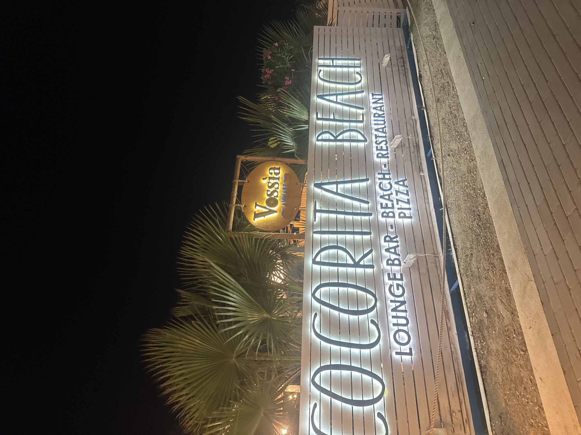 The image shows a nighttime exterior view of "Coccorita Beach" and "Vossia" establishments, likely located in Isola delle Femmine, Italy. A large, illuminated white wooden fence displays the name "Coccorita Beach" in large, glowing white letters. Below this, smaller glowing blue letters spell out "LOUNGE BAR - BEACH - RESTAURANT PIZZA". A circular sign with the name "Vossia" in glowing yellow letters is visible above and to the right of the "Coccorita Beach" sign. Palm fronds are prominent in the background, illuminated by artificial light. The ground in front of the fence appears to be a mix of gravel and paving stones. The overall scene suggests a beachside venue at night.