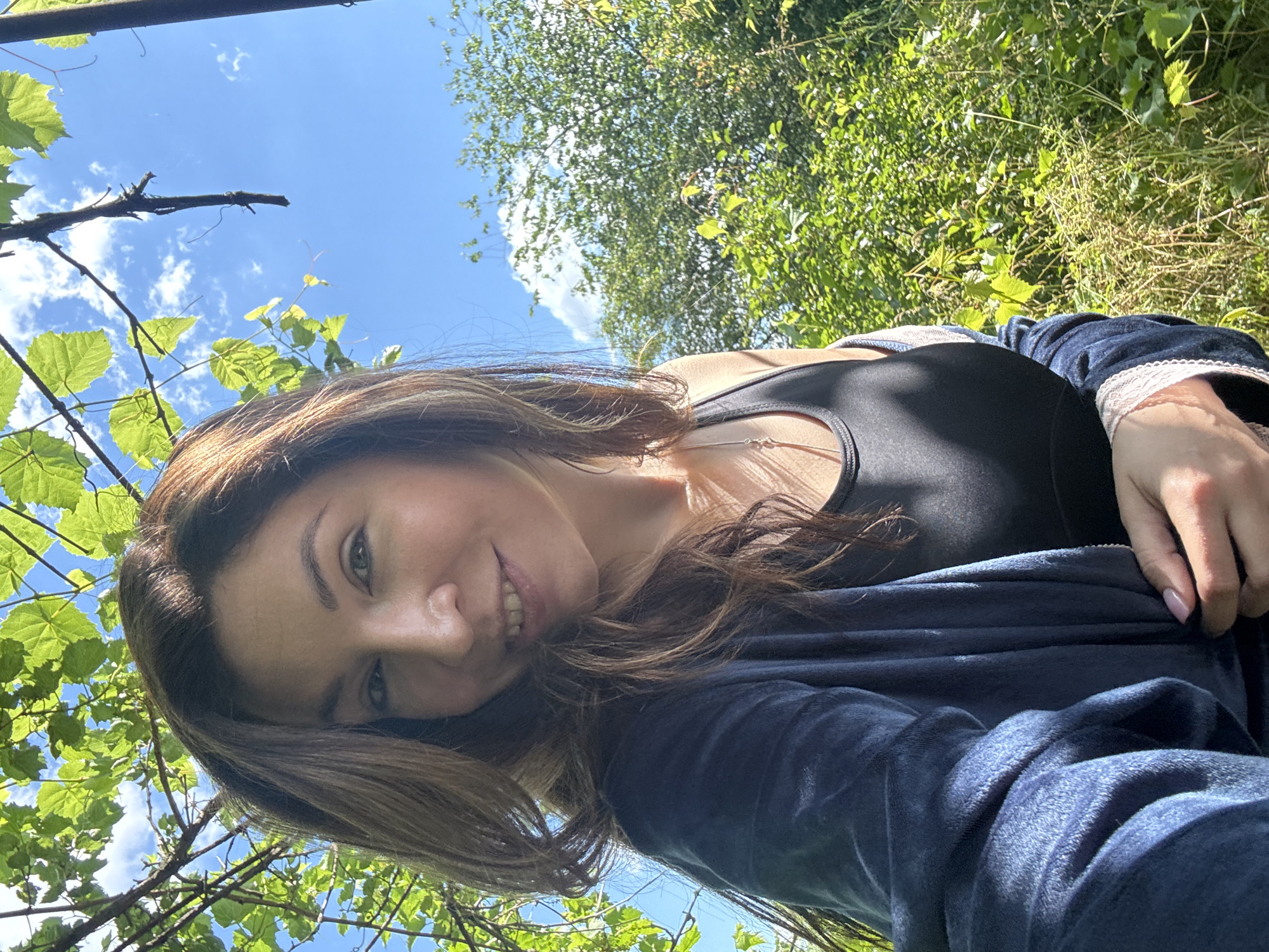A woman with long, brown hair and fair skin is captured in a selfie taken outdoors. She is wearing a dark blue velour robe over a black tank top. Her right hand is visible, with light pink nail polish on her fingernails, as she adjusts the robe. The background consists of lush green foliage, with sunlight dappling through the leaves. The sky is bright blue with scattered white clouds. The scene appears to be a sunny day in a natural setting, possibly a garden or park, in Lozuvatka, Ukraine.