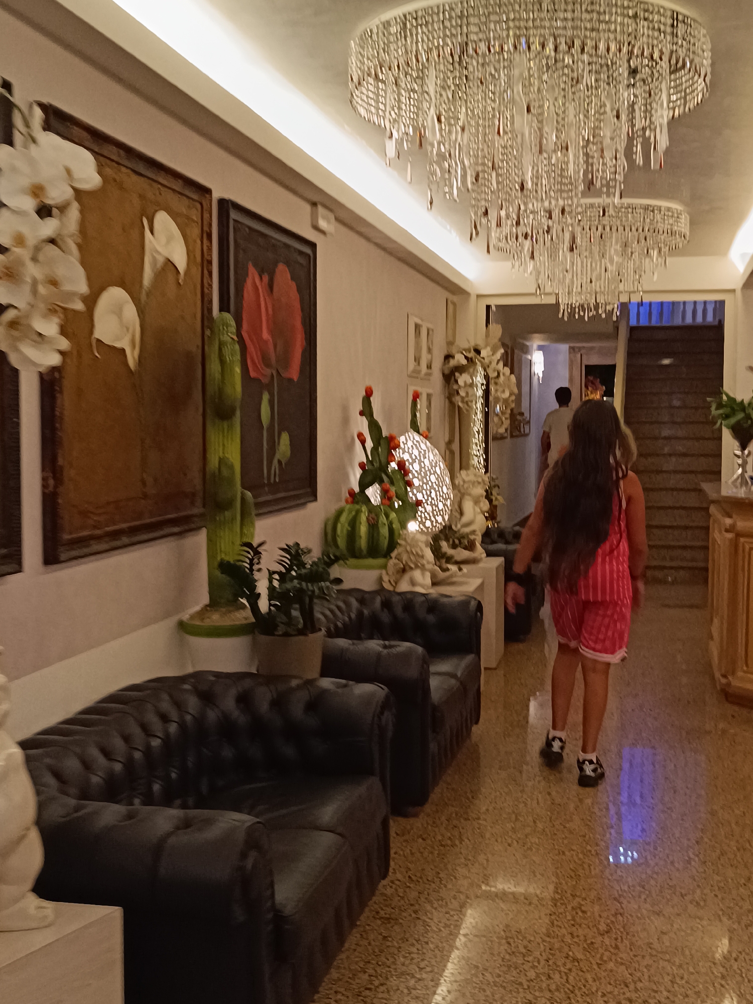 The image shows the interior of a building, likely a hotel lobby or a similar public space, with elements suggesting it's located in Gatteo, Italy. A young girl with long dark hair, wearing a pink striped outfit and white and black sneakers, is walking away from the camera down a hallway. She is in the foreground, and behind her, a person in a light-colored shirt is walking in the same direction.
The environment features a long hallway with a polished terrazzo floor that reflects the overhead lighting. Two large, dark tufted leather sofas are visible on the left side of the hallway. On the wall to the left, there are several framed artworks. One prominent artwork depicts white orchids, and another shows two red poppy-like flowers. Between these artworks and further down the wall, there are decorative potted plants, including a tall, segmented green cactus-like sculpture and a smaller potted succulent. A large, elaborate crystal chandelier hangs from the ceiling, with another smaller one visible further down.
On the right side of the hallway, a grand staircase with wooden steps is visible, leading upwards. A wooden cabinet with ornate carvings is situated to the right of the staircase. The lighting is warm and ambient, emanating from the ceiling fixtures and the chandeliers. The overall impression is one of a well-decorated and well-lit indoor space, possibly a hotel in Gatteo, Italy, given the context.