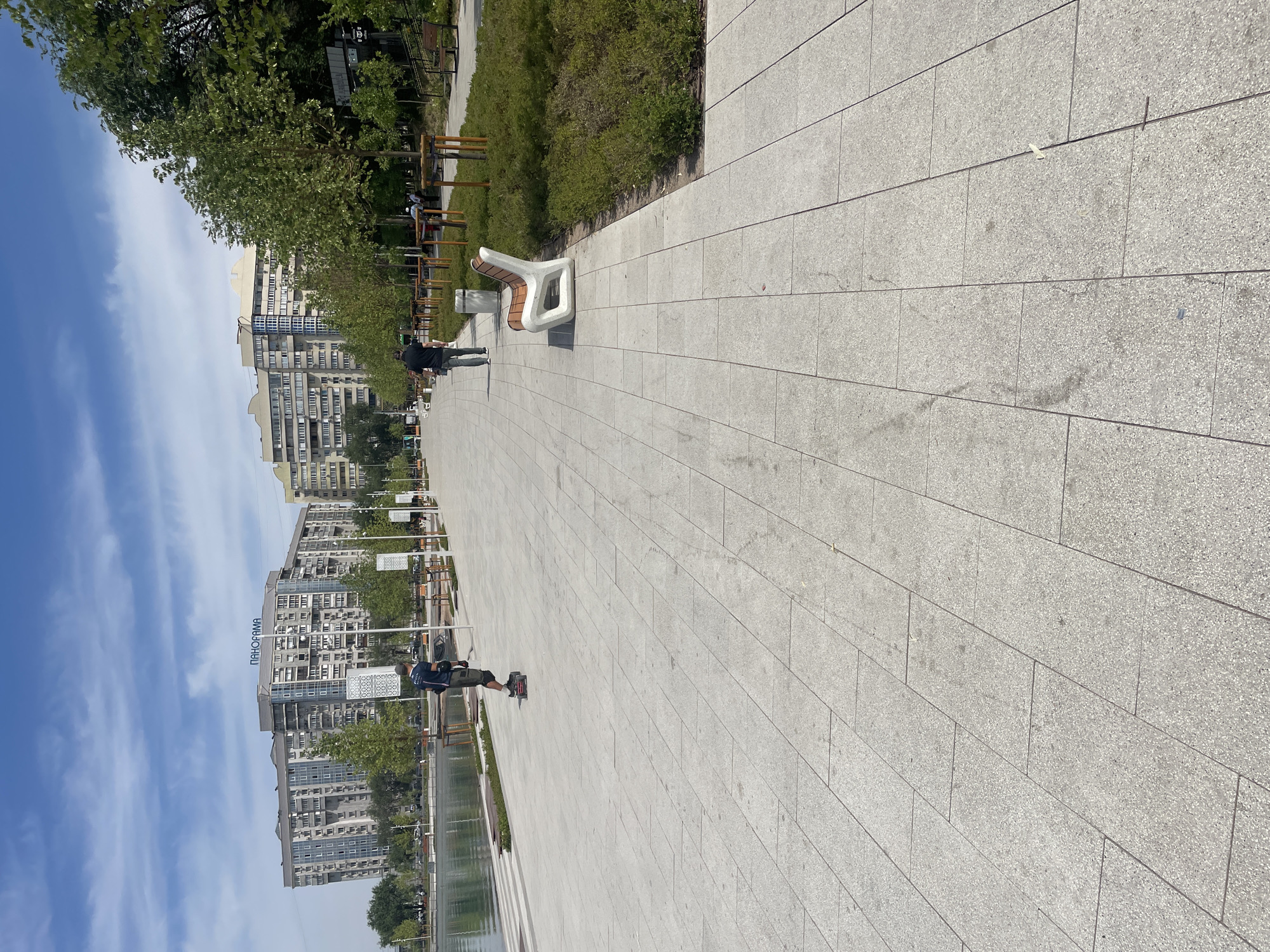 This image depicts a sunny day along the embankment of the Esentai River in Almaty, Kazakhstan. The foreground is dominated by a wide, light-colored tiled pedestrian walkway. To the left, the calm, reflective surface of the river borders the walkway, separated by a low concrete barrier.
On the walkway, a person on an electric skateboard is moving away from the camera. Further down the path, another individual is walking towards the camera, dressed in dark clothing. In the middle ground, several modern, minimalist benches with white frames and wooden slats are strategically placed. These are interspersed with young trees planted in manicured green areas.
The background features a row of multi-story apartment buildings, some with the prominent blue "PANORAMA" sign visible on one of them, indicating the area's urban setting. The sky is a clear blue with scattered white clouds. The overall scene suggests a well-maintained public recreational space within a city environment.