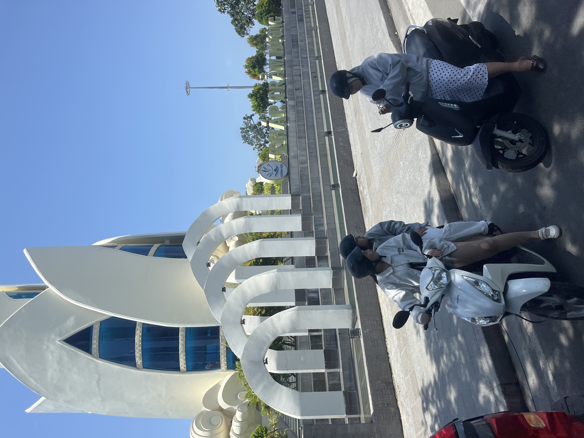 In Nha Trang, Vietnam, two women on scooters are parked on a paved road. The woman on the left, wearing a light-colored jacket and black helmet, sits on a white scooter, while the woman on the right, in a gray hoodie and black helmet, is on a black scooter, adjusting her glasses. Behind them, a large white lotus-shaped building dominates the background, with blue-tinted windows. A row of arched white structures stands in front of the building, leading to a set of tiered stairs with large golden lettering that reads "THAP TRAM HUONG" and a logo for "KHANH HOA". The sky is clear and blue, and trees are visible in the distance. The overall scene suggests a bright, sunny day at a landmark in the city.