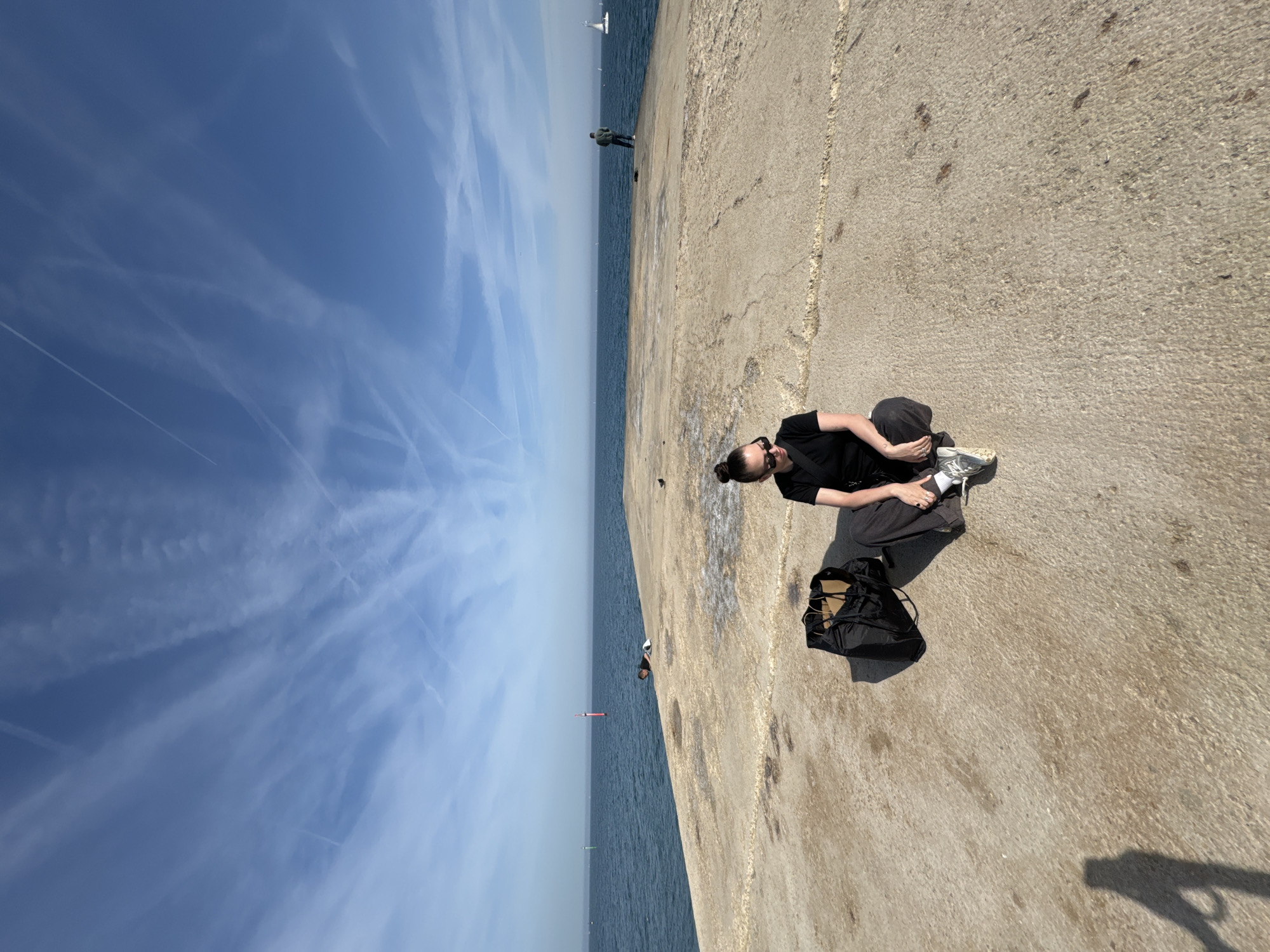 A horizontal shot depicts a seated person with a backpack along a concrete pier. The person, wearing dark clothing and sunglasses, has their hair in a bun. Their legs are bent, and they appear to be focused on something in their lap. Beside them is a black backpack.
The pier extends into the sea, with the sky and the sea divided by a distant horizon. Other people can be seen standing further along the pier. The sky is mostly blue, streaked with cloud patterns. There is a navigational marker in the sea.
The setting is Barcelona, Spain, specifically the seawall near Port Vell.