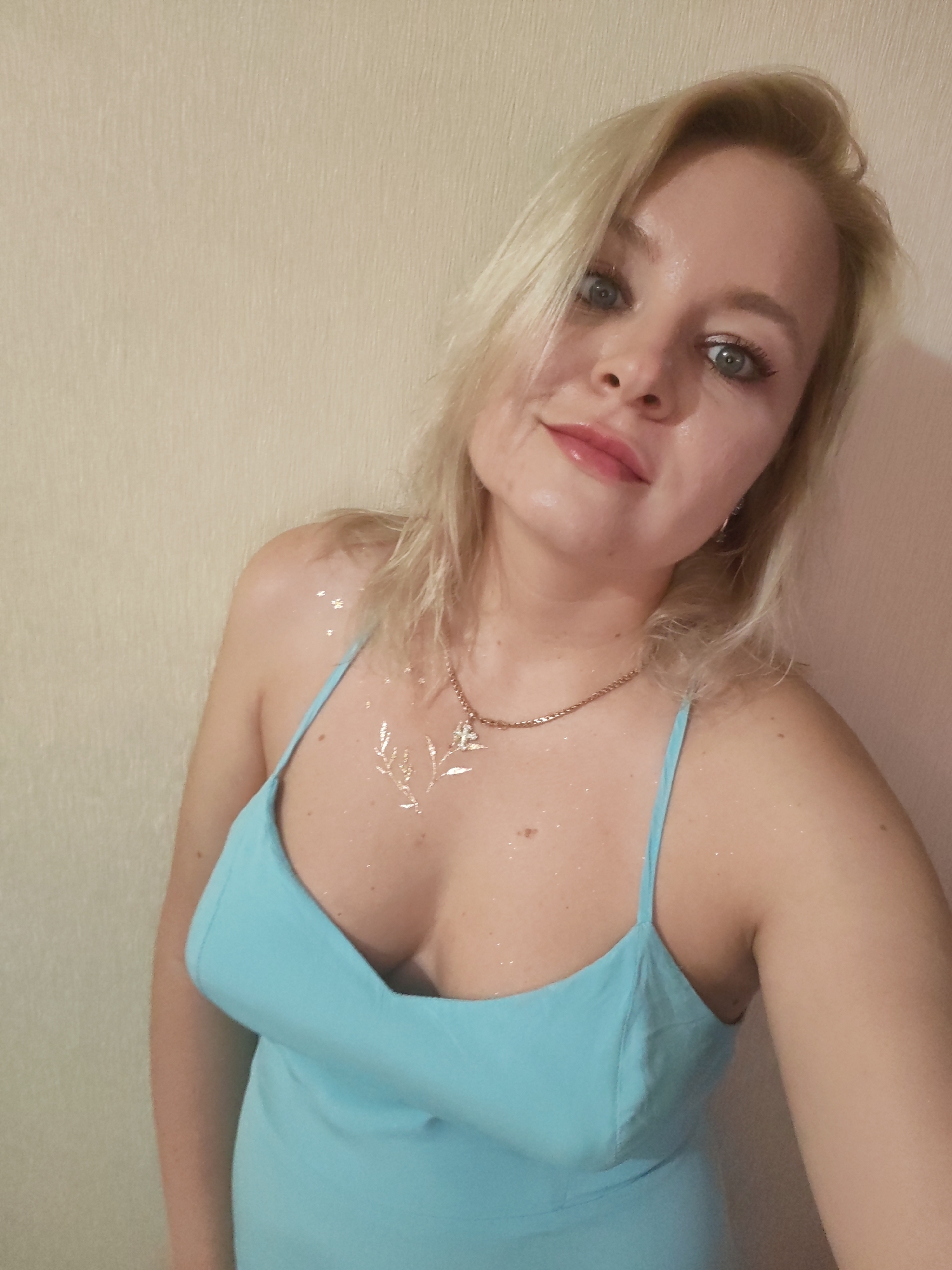 A blonde woman with blue eyes and pink lipstick is shown in a close-up shot. She is wearing a light blue slip dress and a gold chain necklace. Her chest is decorated with glitter, and she has a small branch design also made of glitter on her collarbone. The background is a plain, textured wall. The image does not contain any information to suggest it was taken in Izhevsk, Russia.