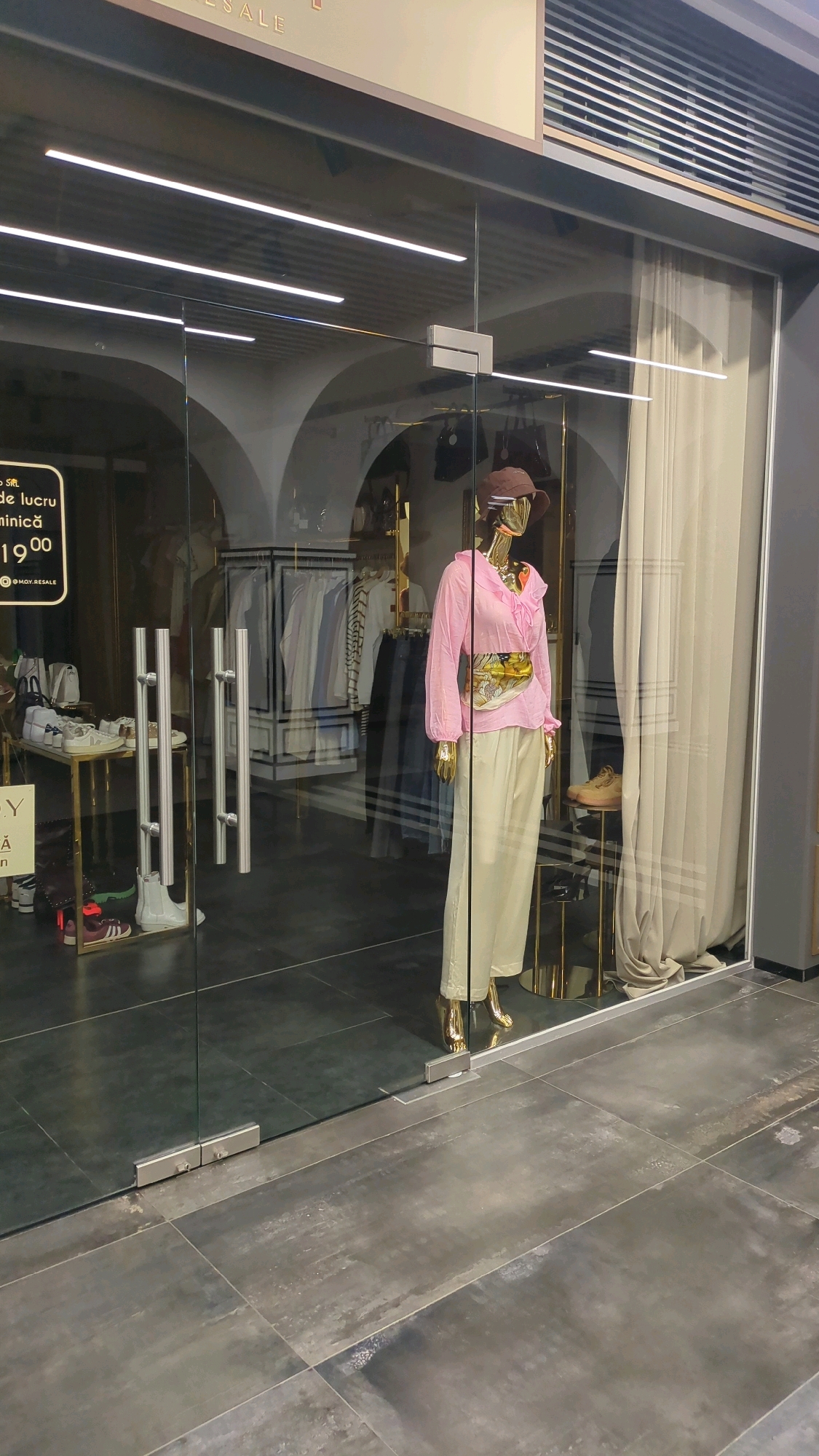 The image shows the storefront of a boutique in Chișinău, Moldova. The boutique features a large glass window and double glass doors with polished metal handles. Inside, a mannequin is displayed wearing a pink blouse, light-colored wide-leg pants, a gold belt, and a brown hat. The store interior includes clothing racks with various garments, a display table with shoes, and a tall gold pedestal displaying a sneaker. The flooring inside appears to be tiled, while the exterior ground is a darker, textured material. A sign on the left side of the window is partially visible, displaying text in Romanian. The overall impression is one of a modern and stylish retail space.