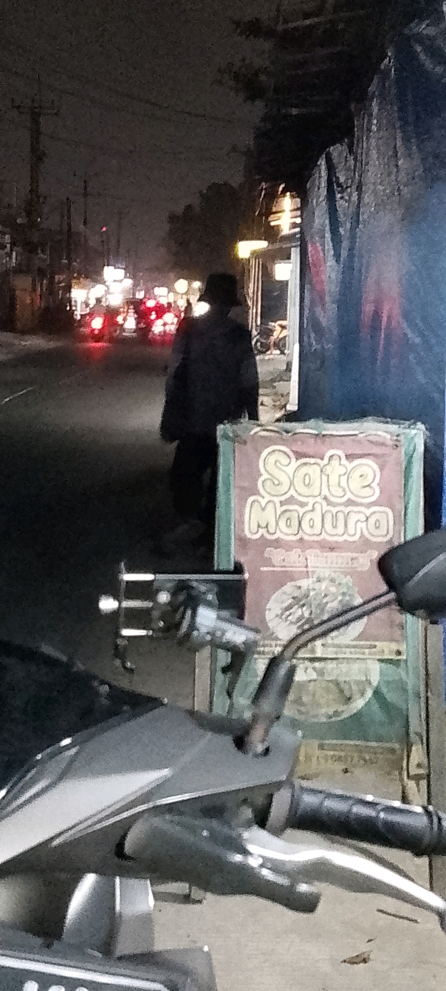 The image captures a nighttime scene on a street, likely in Satriamekar, Indonesia. In the foreground, the handlebars and front portion of a motorcycle are visible, with a silver-grey body and black grips. To the right, a sign is displayed, featuring text that reads "Sate Madura" in large, stylized letters. Below this, there's smaller text and an image of what appears to be a bowl of satay. The sign is bordered by green and pink. In the mid-ground, a person wearing a dark jacket and a hat walks away from the camera, heading towards a street with traffic. The street is illuminated by the headlights and taillights of vehicles, creating streaks of light. Several motorcycles with illuminated taillights are visible in the distance. The background shows buildings, utility poles, and trees under the dark, possibly cloudy, night sky. The overall impression is that of a bustling street at night, possibly a commercial area.