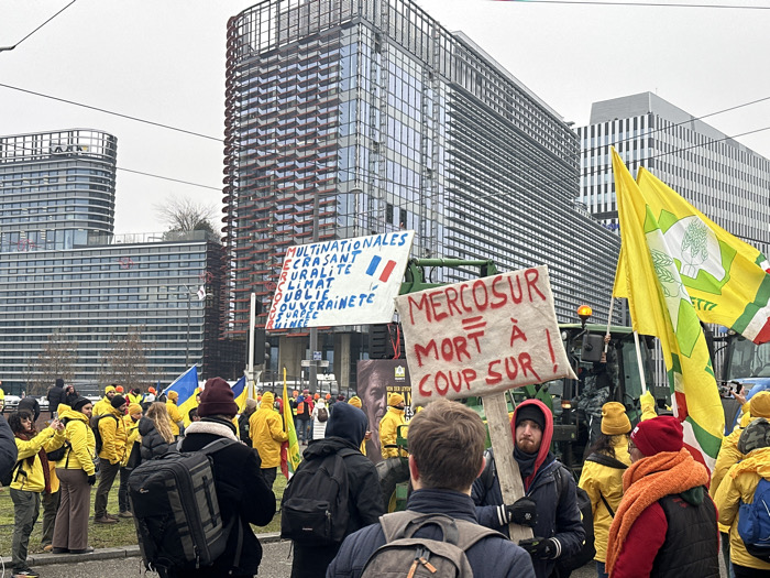 The image depicts an outdoor protest occurring on an overcast day in Strasbourg, France, located in the vicinity of the European Parliament. The background features the modern, multi-story glass and steel architecture of the European Parliament's Louise Weiss building, recognizable by its curvilinear design and red-accented facade, alongside other contemporary structures.
In the foreground and midground, a crowd of individuals is gathered, predominantly adults, many wearing bright yellow jackets and hats. A green tractor is partially visible in the midground, positioned behind a protest sign.
Two primary protest signs are visible. A large white banner, featuring a French flag motif, is displayed on the left with text in French that reads: "MULTINATIONALES ÉCRASANT RURALITÉ CLIMAT OUBLIÉ SOUVERAINETÉ USURPÉE RUINÉE" (Multinationals crushing rurality, forgotten climate, usurped/ruined sovereignty). In the foreground, center-right, a male individual with a beard and red hat holds a smaller, rectangular cardboard sign with red lettering stating: "MERCOSUR = MORT À COUP SÛR !" (Mercosur = Death for sure!).
Several yellow flags, each bearing a green stylized agricultural emblem, are prominently displayed throughout the crowd, alongside at least one blue and yellow Ukrainian flag. Participants are standing, observing, and holding signs, with some individuals appearing to record the event with mobile phones. The overall scene indicates an organized public demonstration.