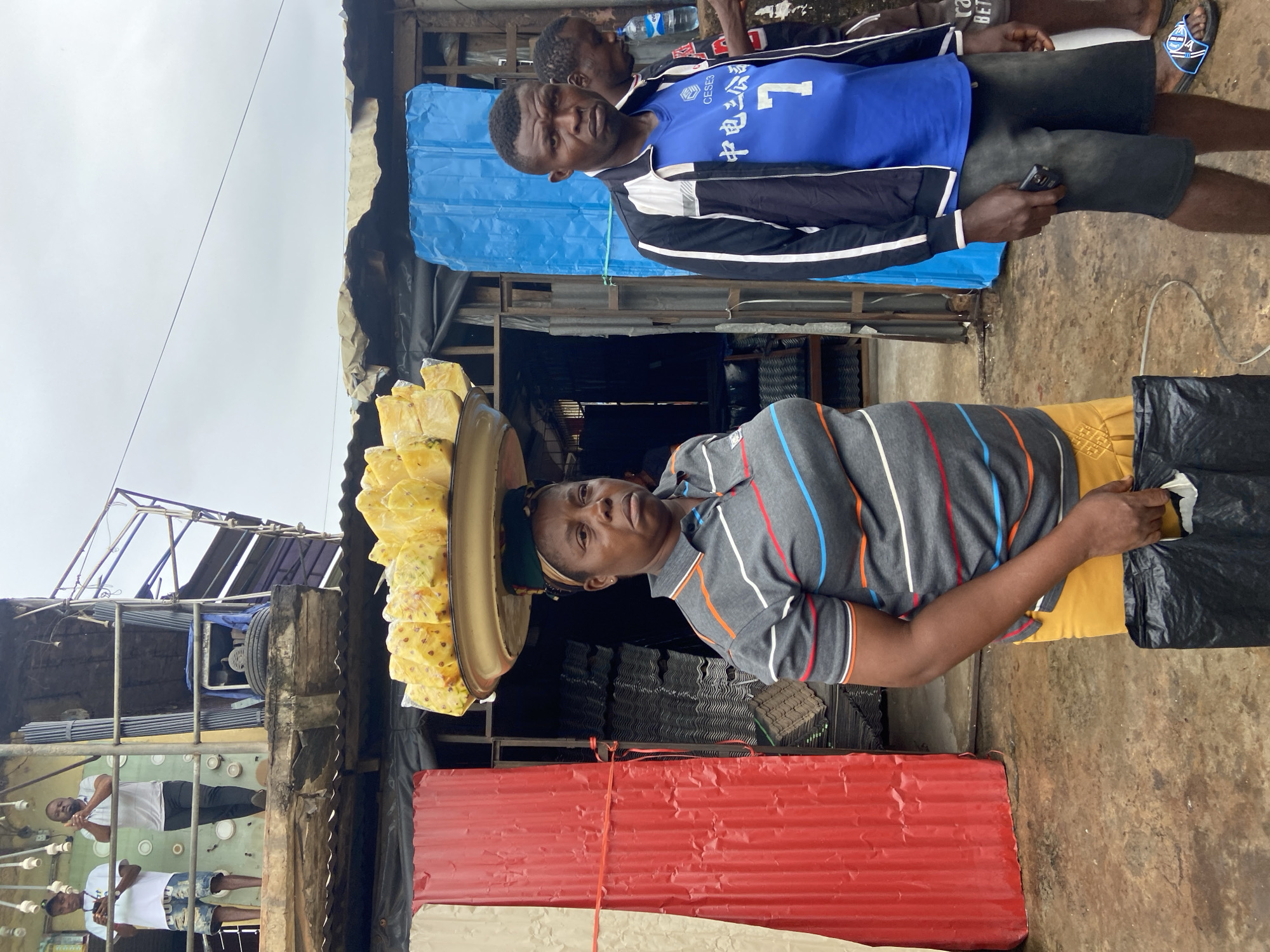 In Ogbunike, Nigeria, a woman with dark skin and a serious expression stands in the foreground, carrying a large tray of cut pineapple on her head. She is wearing a gray t-shirt with horizontal stripes of orange, white, and blue, and a yellow skirt. Her right hand is holding a black plastic bag. To her right, a man with dark skin and short hair looks directly at the camera. He is wearing a blue t-shirt with Chinese characters and the number "7" printed on it, a black and white jacket, and dark shorts. Behind them, a market stall is visible with corrugated metal walls painted red and cream on the left, and a bright blue tarp on the right. In the background, other individuals are partially visible, and there is evidence of makeshift structures and overhead wires. The ground appears to be a rough, dirt surface.