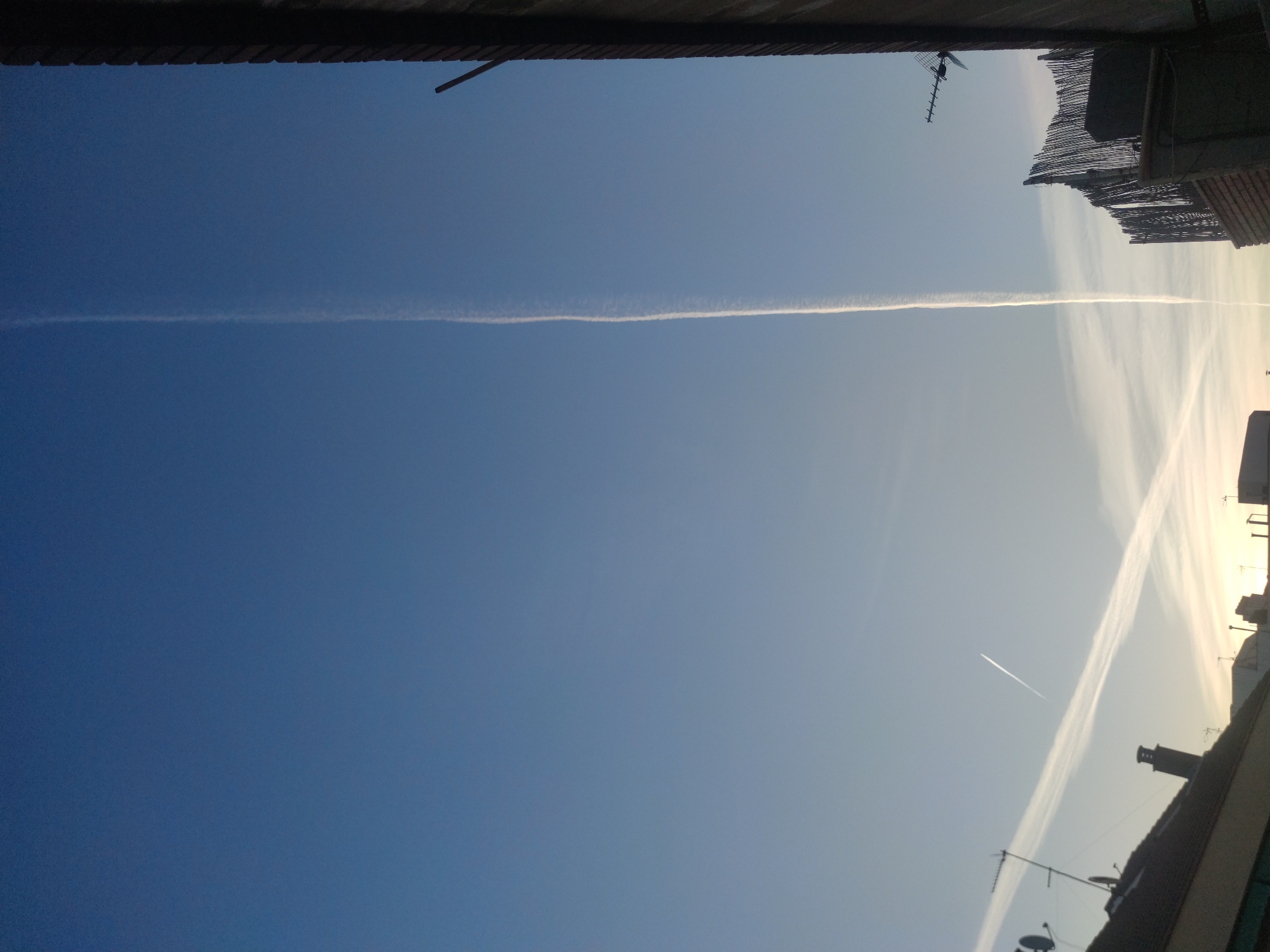 The image depicts a sky view from a city, possibly from a rooftop or a window. The sky is mostly clear with varying shades of blue, and contrails are visible, suggesting aircraft activity. The upper and lower edges of the image show portions of buildings, including rooftops, antennas, and chimneys. The city location is unspecified.