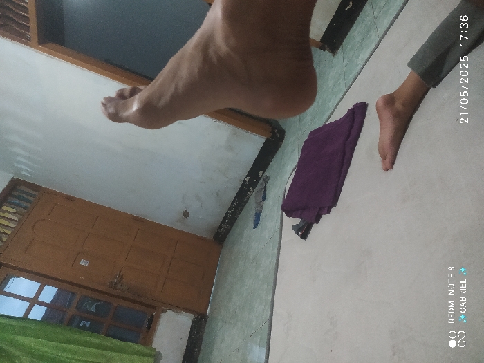 The image shows the interior of a home in Kebumen, Indonesia, with a white tiled floor. A person is lying on a rug, with their right foot raised towards the ceiling, and the bottom of their right leg is in view. There is a wooden door to the left of the image, and a wall to the right. A purple towel lies on the rug, and a shadow of a small object can be seen near it. The timestamp on the bottom right corner of the image indicates the date and time as 21/05/2025 17:36.