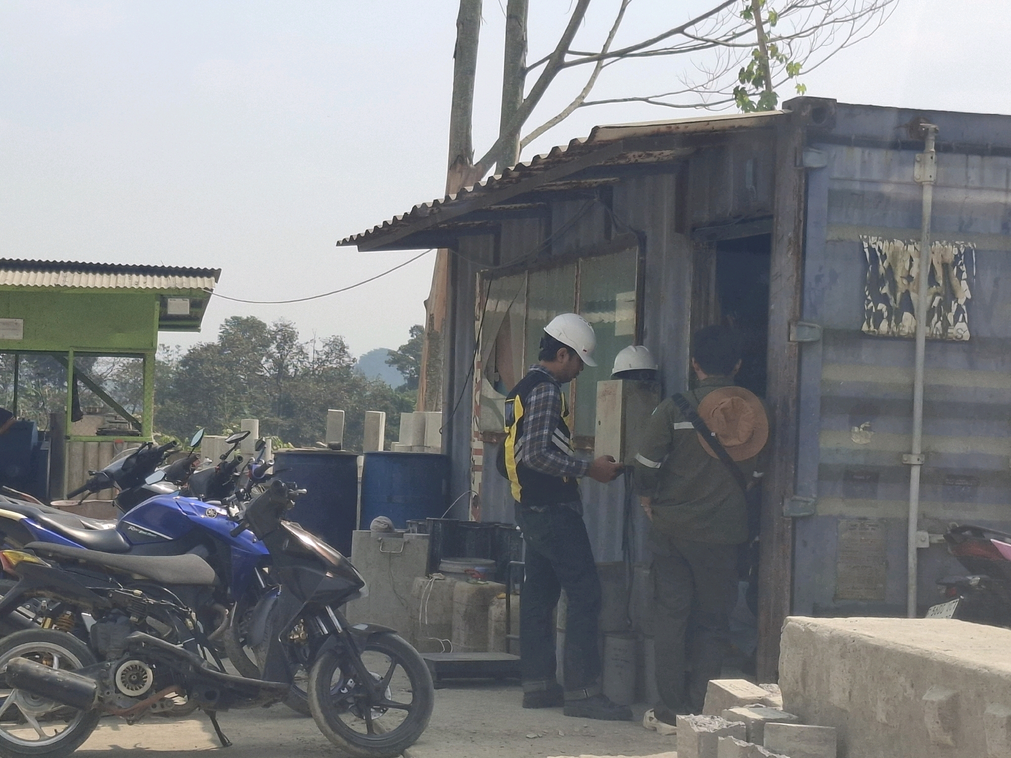 The image depicts an outdoor scene, likely in Cijangkar, Indonesia, with a focus on a blue shipping container that has been converted into a small structure with a corrugated metal awning. Three individuals are present. The first, wearing a white hard hat and a plaid shirt with a yellow safety vest, stands to the left of the container entrance, holding a mobile device. To the right of the entrance, a second individual, also wearing a white hard hat and a dark green work uniform, stands with their back to the camera, wearing a tan hat. A third individual, partially visible inside the container, appears to be interacting with the person in the green uniform. In the foreground, several motorcycles are parked, including a blue and black Yamaha motorcycle. Behind the container, large blue drums and stacks of concrete blocks are visible. The environment appears to be an industrial or storage area, with trees and a hazy sky in the background.
