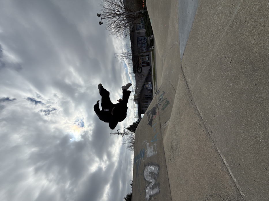 The image displays an outdoor concrete skatepark in Shumen, Bulgaria, located adjacent to a commercial building and city park area. A single individual is captured in mid-air, silhouetted against a cloudy sky, positioned in the upper central portion of the frame. The subject wears dark, loose-fitting attire and light-colored athletic footwear, with limbs bent, indicating a dynamic jump or aerial maneuver.
The skatepark occupies the lower half of the image, consisting of various grey concrete ramps, ledges, and flat sections. Faded graffiti, including a prominent letter "M," is visible on some of the sloped concrete surfaces. The sky is overcast with dense clouds, through which the sun is partially visible in the upper left, surrounded by a faint iridescent halo or sun dog effect.
In the background, behind the skatepark, a two-story building with a flat roof, large windows, and an external staircase is visible. Signage on the building includes the text "PIMI." Bare deciduous trees are scattered across the background, particularly on the right, where a tall streetlight with multiple fixtures is also present. To the far left background, a metal electricity pylon is visible against the sky. The overall light suggests an overcast day with the sun partially breaking through.