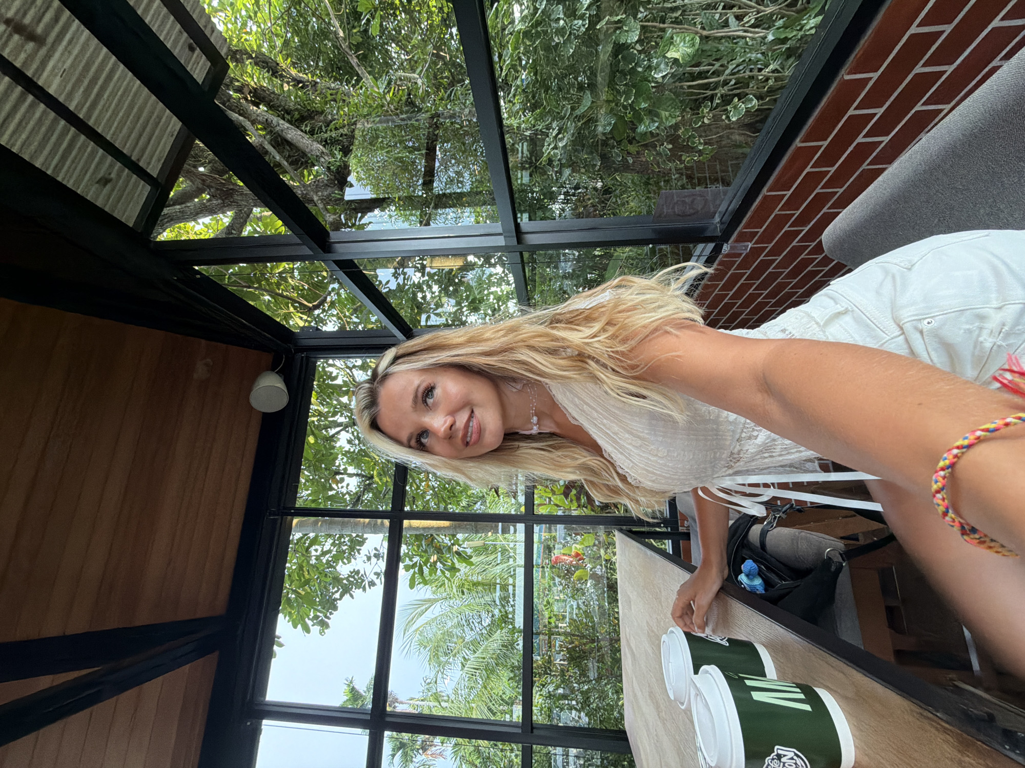 A young Caucasian woman with blonde hair is seated at a wooden table in an open-air establishment, possibly a cafe or restaurant, in Sakhu, Thailand. She is wearing a white lace top and white shorts. She is holding a smartphone in her right hand, taking a selfie. Two green disposable coffee cups with white lids are placed on the table in front of her. Through the large glass windows, lush green trees and tropical foliage are visible, suggesting an outdoor setting within a natural environment. The interior features exposed wooden beams and brickwork, indicating a rustic or modern-tropical architectural style. The overall ambiance is casual and relaxed, with natural light filtering through the greenery.
