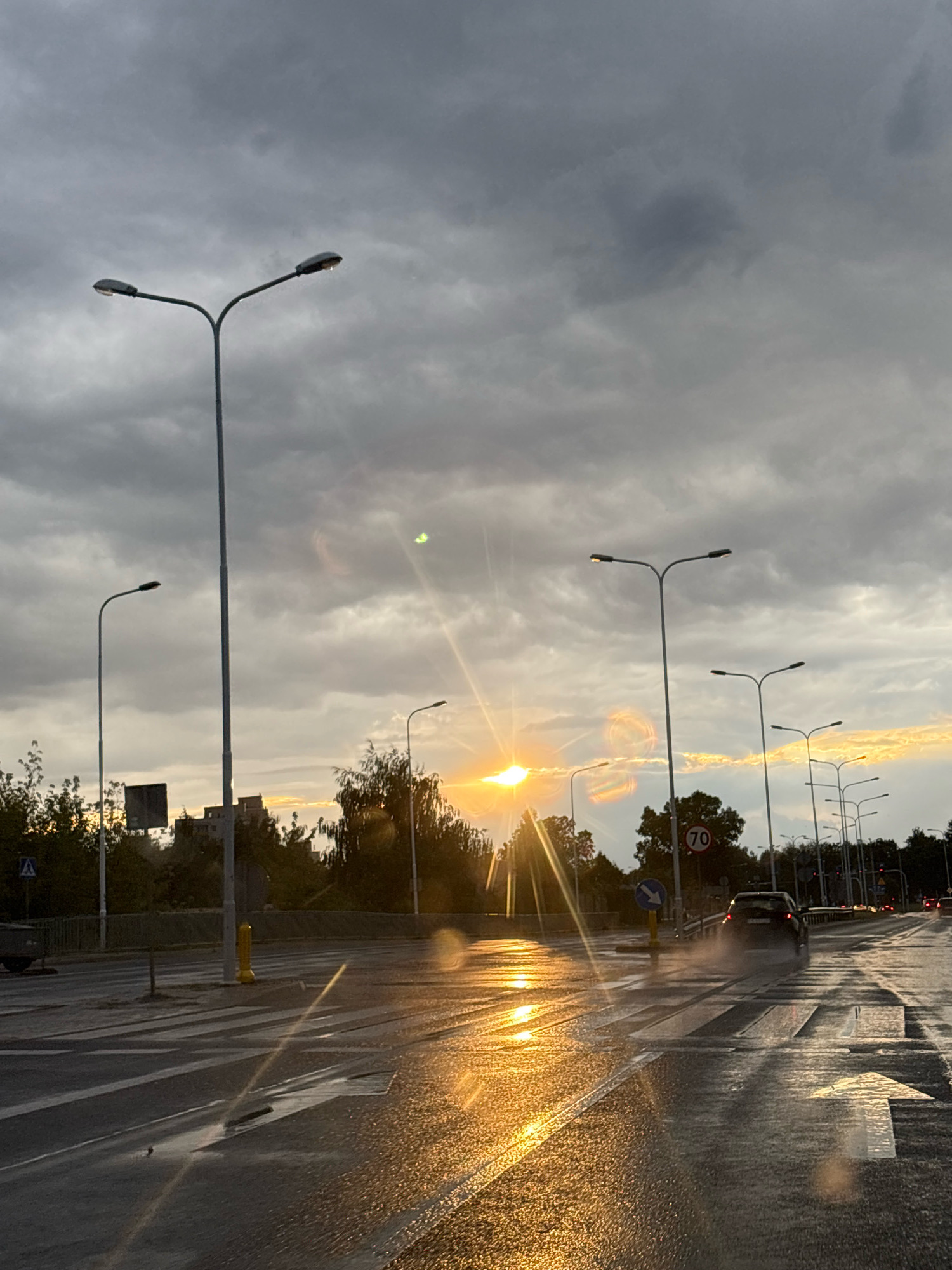 The image captures a street scene in Lublin, Poland, during what appears to be a rainy or post-rain evening. The sky is heavily overcast with dramatic, dark clouds, but a bright, low-hanging sun breaks through, casting a strong, golden reflection on the wet asphalt. The wet road surface creates a mirror-like effect, highlighting the sun's rays and the illuminated markings of a pedestrian crossing.
Several streetlights are visible, lining the road and rising into the cloudy sky. One prominent streetlight in the foreground has a distinctive Y-shaped design with two lamps. Further down the road, a line of similar streetlights stretches into the distance.
A dark-colored car is seen driving away from the viewer, its tires kicking up spray from the wet road. A speed limit sign indicating "70" is mounted on a pole beside the road, as is a blue directional sign with a white arrow. Trees and some low buildings are visible in the background, silhouetted against the brighter sky. The overall atmosphere is one of a moody, wet evening in an urban setting.