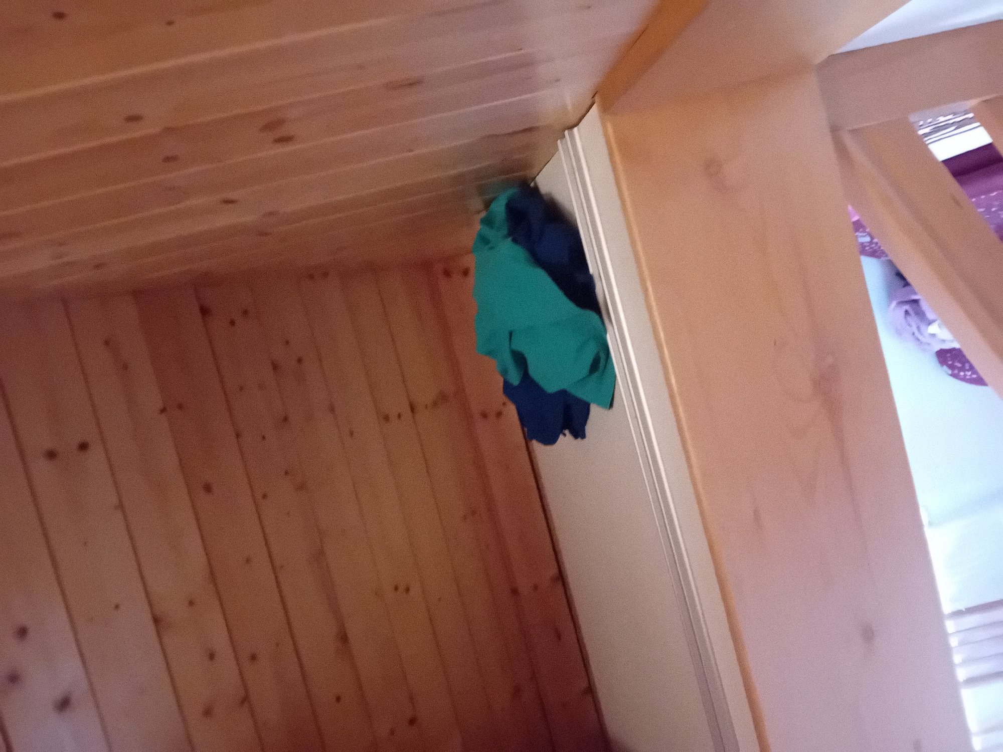A pile of folded blue and teal fabric sits on a white shelf, which is positioned below a wooden sloped ceiling made of horizontal planks. The shelf is at the edge of a wooden loft or mezzanine level, with a light-colored surface extending below it. The wall to the right of the shelf is also made of vertical wooden planks. The scene appears to be indoors, possibly in a residential attic or loft space in Zuberec, Slovakia.