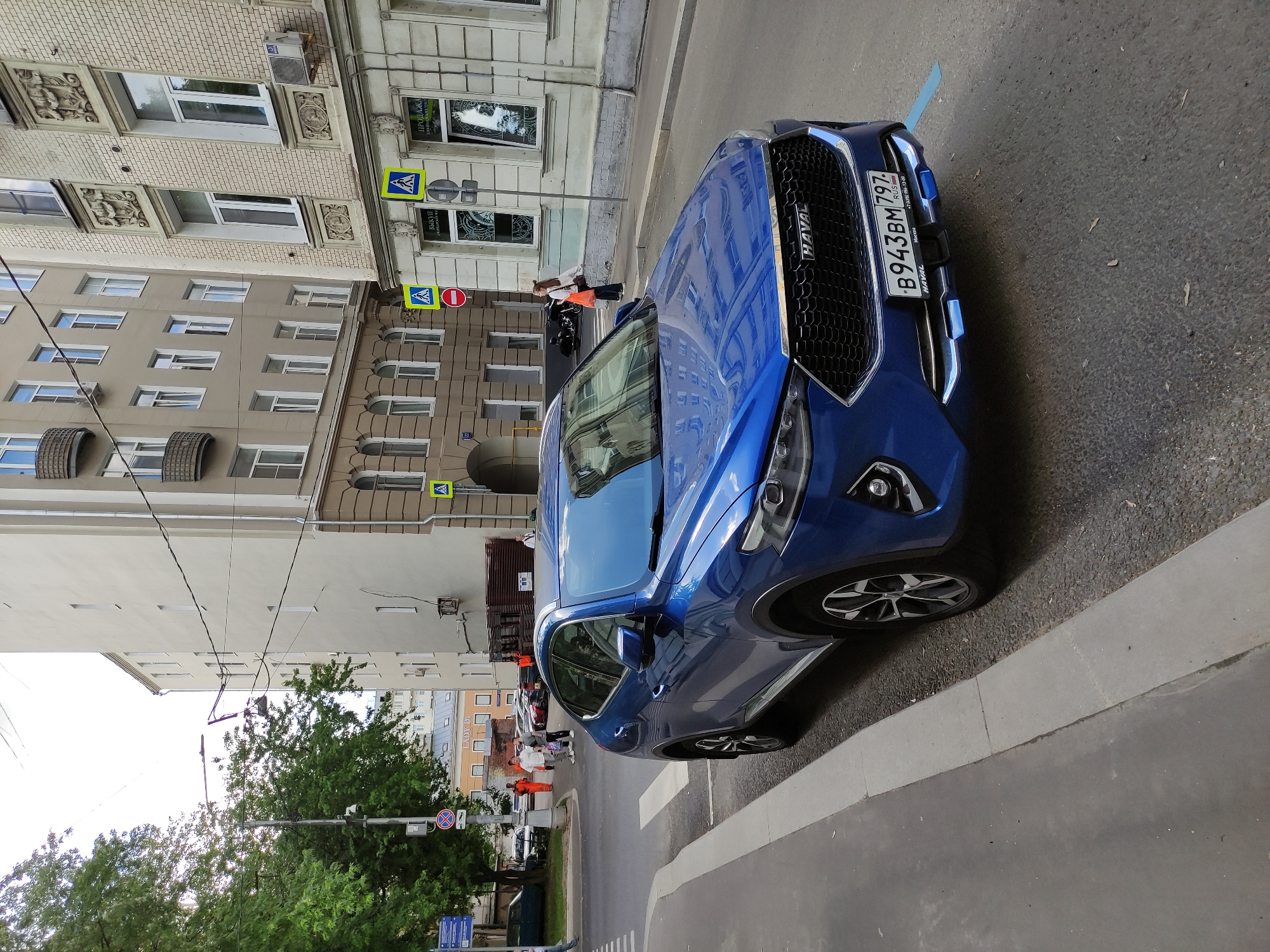 A blue Haval car is parked on the side of a city street in what appears to be Unknown, Unknown. The license plate number is B943BM797. A crosswalk is visible in the background with several pedestrians walking across it. There are buildings on the opposite side of the street, and trees on the near side of the street. The day seems to be overcast.