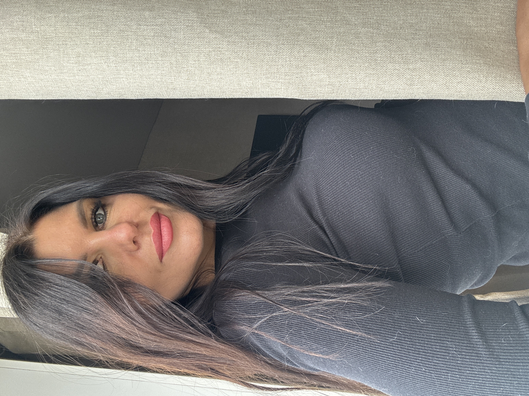 A brunette woman with fair skin and green eyes is seen from a low angle, her face partially obscured by dark hair. She has pink lipstick on. She is wearing a black ribbed sweater. The background includes a light-colored, textured surface, possibly a cushion or upholstery, and a glimpse of a dark, indeterminate space. There is not enough information to confirm that the image was taken in the city of Chaikovsky, Russia.