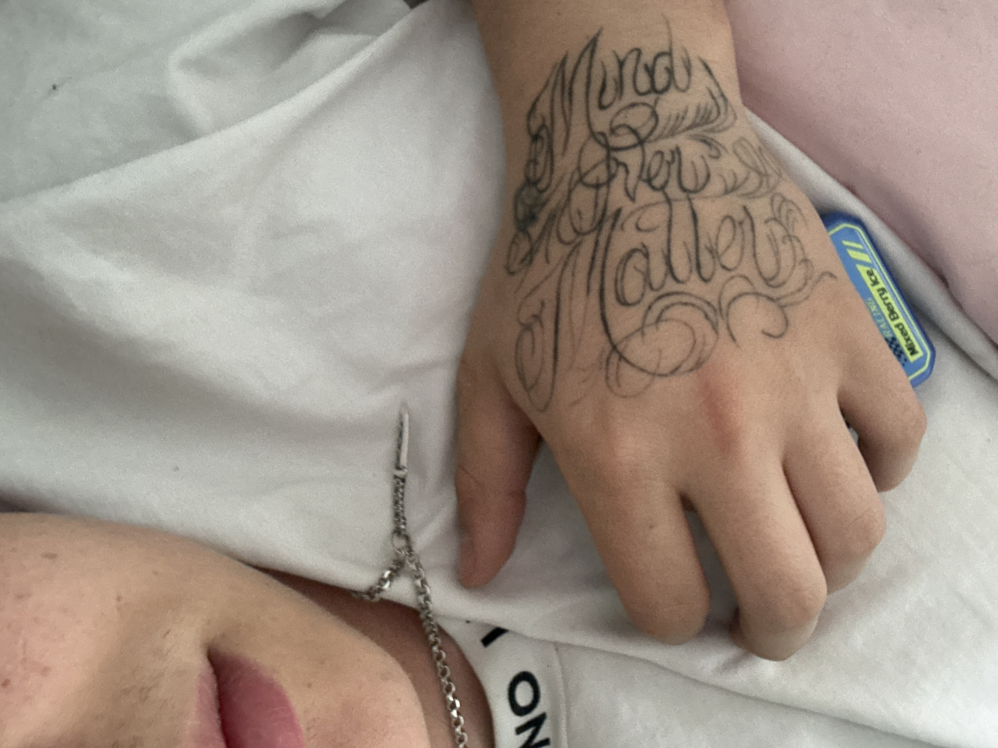 A close-up shot depicts a person lying on a white surface, possibly a bed. The person's left hand, adorned with a black ink tattoo of stylized text that reads "Mind Over Matter," is prominent. A silver chain, possibly a necklace, is visible next to the hand. Part of the person's face, including their mouth with pink lipstick, is also in view. A blue and yellow item, possibly a medication or lip balm, rests near the hand. The location is unknown.