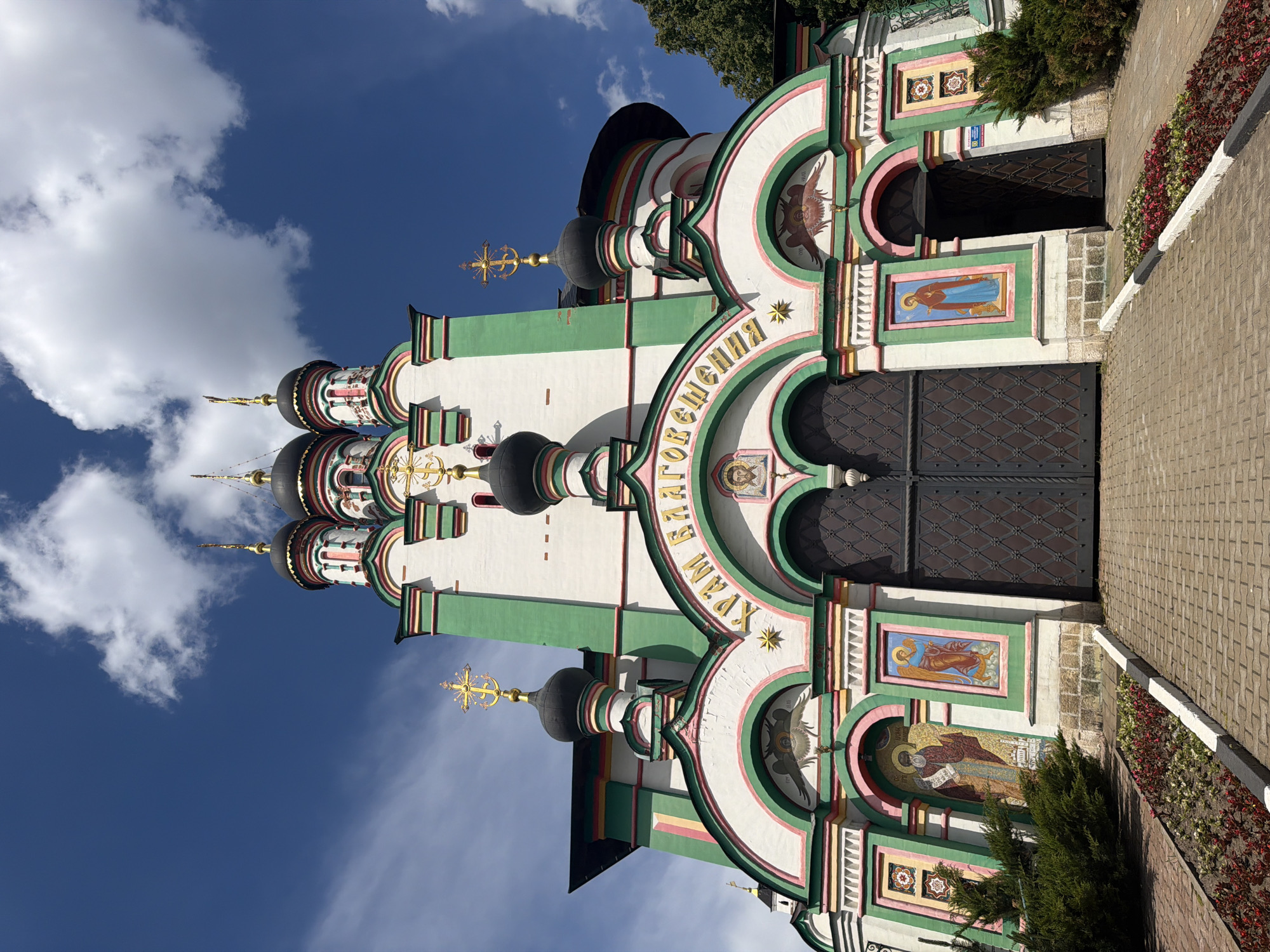The image displays the vibrant and intricately detailed facade of an Eastern Orthodox church in Pavlovskaya Sloboda, Russia. The church exterior features white walls with ornate decorative elements in green, pink, red, and gold. Several dark, onion-shaped domes (cupolas), each topped with a golden Orthodox cross, rise against a bright blue sky dotted with fluffy white clouds.
The main entrance consists of large, dark-colored double doors with a geometric pattern, situated beneath a grand archway. A smaller, recessed entrance is visible to the right. The facade is richly adorned with religious art, including a large mosaic depicting a male saint with a halo and a scroll, a smaller painting of a female saint in blue robes (likely the Virgin Mary), and an emblem resembling an eagle or phoenix above the main doors. A golden star is also visible on the facade.
Along the right side of the church, a paved pathway of bricks or cobblestones is present, bordered by a white curb and landscaped with red and green ground cover plants and a small evergreen bush. The clear sky and bright illumination indicate it is daytime, likely midday or early afternoon.
Prominently displayed in gold Cyrillic text on a green archway above the main entrance are the words "ХРАМ БЛАГОВЕЩЕНИЯ," which translates to "Temple of the Annunciation" or "Church of the Annunciation." The scene captures the architectural grandeur and spiritual significance of the building, with no specific activity or people depicted.