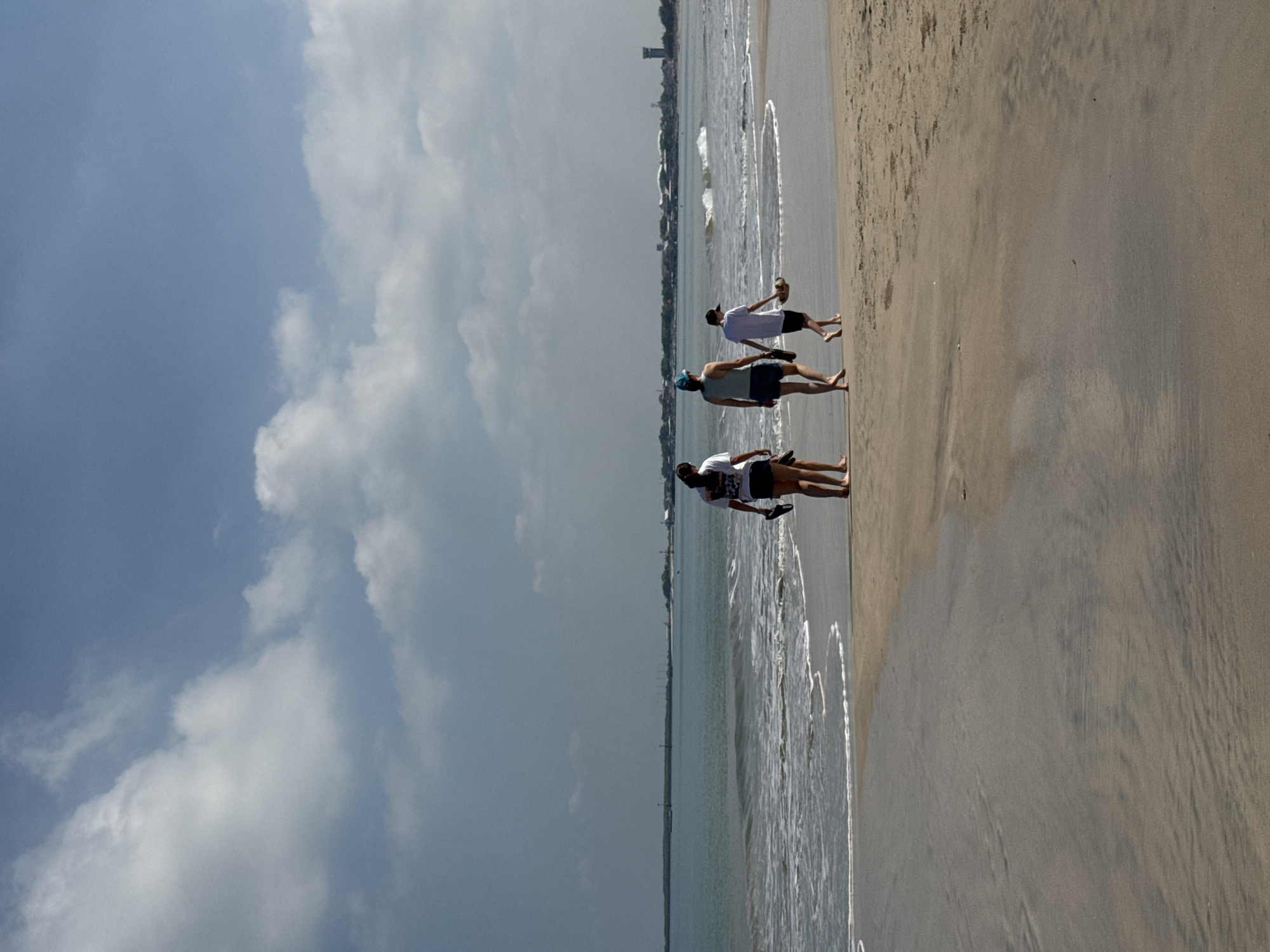 This image depicts a sunny day on a beach in Kuta Selatan, Indonesia. The scene is dominated by a wide stretch of sandy shore meeting the ocean, with a partly cloudy sky above.
Three individuals, appearing to be young adults, are walking along the water's edge. They are casually dressed in t-shirts and shorts, with at least two of them barefoot in the shallow water. The person in the foreground wears a white t-shirt with some illegible dark text on the back. The next person in line wears a light-colored cap, a white t-shirt, and blue shorts. The third person, furthest from the viewer, is holding what looks like a coconut or another tropical fruit, suggesting a leisurely activity.
The beach features light brown sand with subtle patterns left by the receding tide. Gentle waves are breaking along the shoreline, creating white foam. In the background, across the expanse of water, a distant coastline is visible, dotted with buildings and what appears to be a tall structure or tower, indicating a nearby developed area. The sky presents a mix of clear blue and soft white clouds, with some darker cloud formations closer to the horizon. The bright light suggests it is daytime, likely late morning or early afternoon. The overall atmosphere conveyed is one of relaxation and enjoyment in a tropical coastal setting. No decipherable text is clearly visible on any objects or clothing.