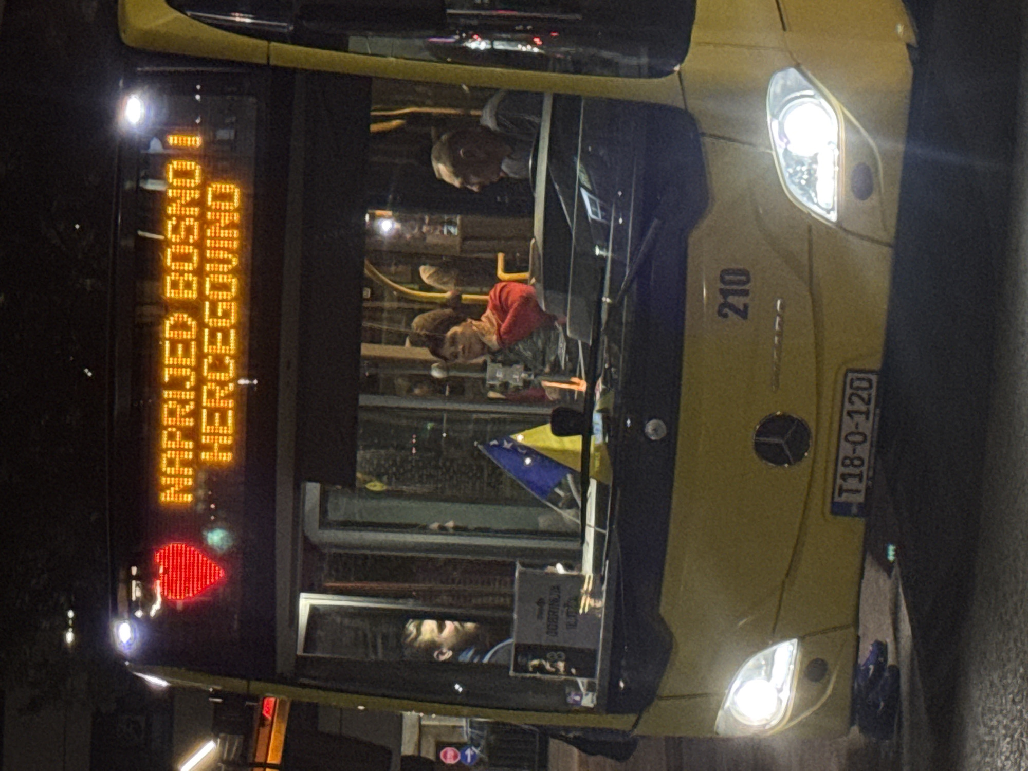 The image shows the front of a yellow Mercedes-Benz bus at night. The bus has its headlights on. Above the windshield, an illuminated sign displays a red heart followed by the text "NAPRIJED BOSNO I HERCEGOVINO" in orange LED letters. Inside the bus, passengers are visible through the windows. A Bosnian flag is displayed inside the bus, near the windshield. A license plate on the front of the bus reads "T18-0-120". The bus appears to be at a bus stop or station, with a traffic light visible in the background to the left. Based on the context of the text on the bus, the location is likely in Ilidža, Bosnia and Herzegovina.