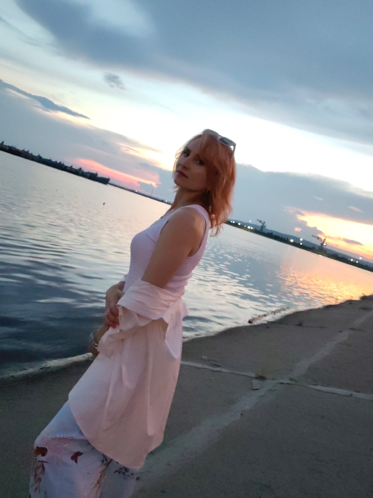 A woman with reddish-brown hair stands on a paved walkway next to a body of water. She is wearing a white tank top, a light pink garment draped over her shoulders and torso, and light-colored pants with a floral pattern. Sunglasses are perched on her head. The sky above is filled with clouds, and the sun is setting over the water, casting warm colors in the distance. Several industrial structures, including cranes, are visible on the far side of the water. The scene appears to be a waterfront area, potentially a port or industrial zone. The presence of industrial cranes and the general landscape suggest a location like the Kama River embankment near the city of Chaikovsky, Russia, which has a significant industrial presence and is situated on the river.