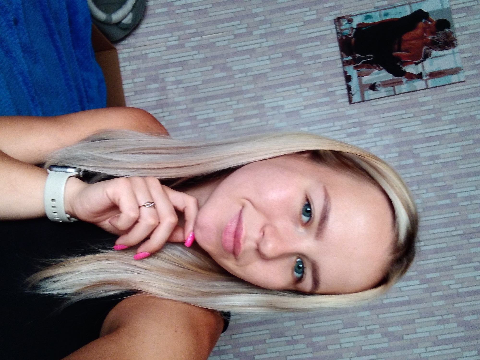 A young woman with blonde hair and blue eyes is captured in a selfie. She is wearing a black top and a smartwatch on her left wrist. Her right hand is resting on her chin, showcasing bright pink fingernails and a ring on her left ring finger. In the background, a wall with a vertical striped pattern and a framed picture of two people holding wine glasses are visible. To the left, a blue blanket is draped over furniture. The image appears to have been taken indoors.