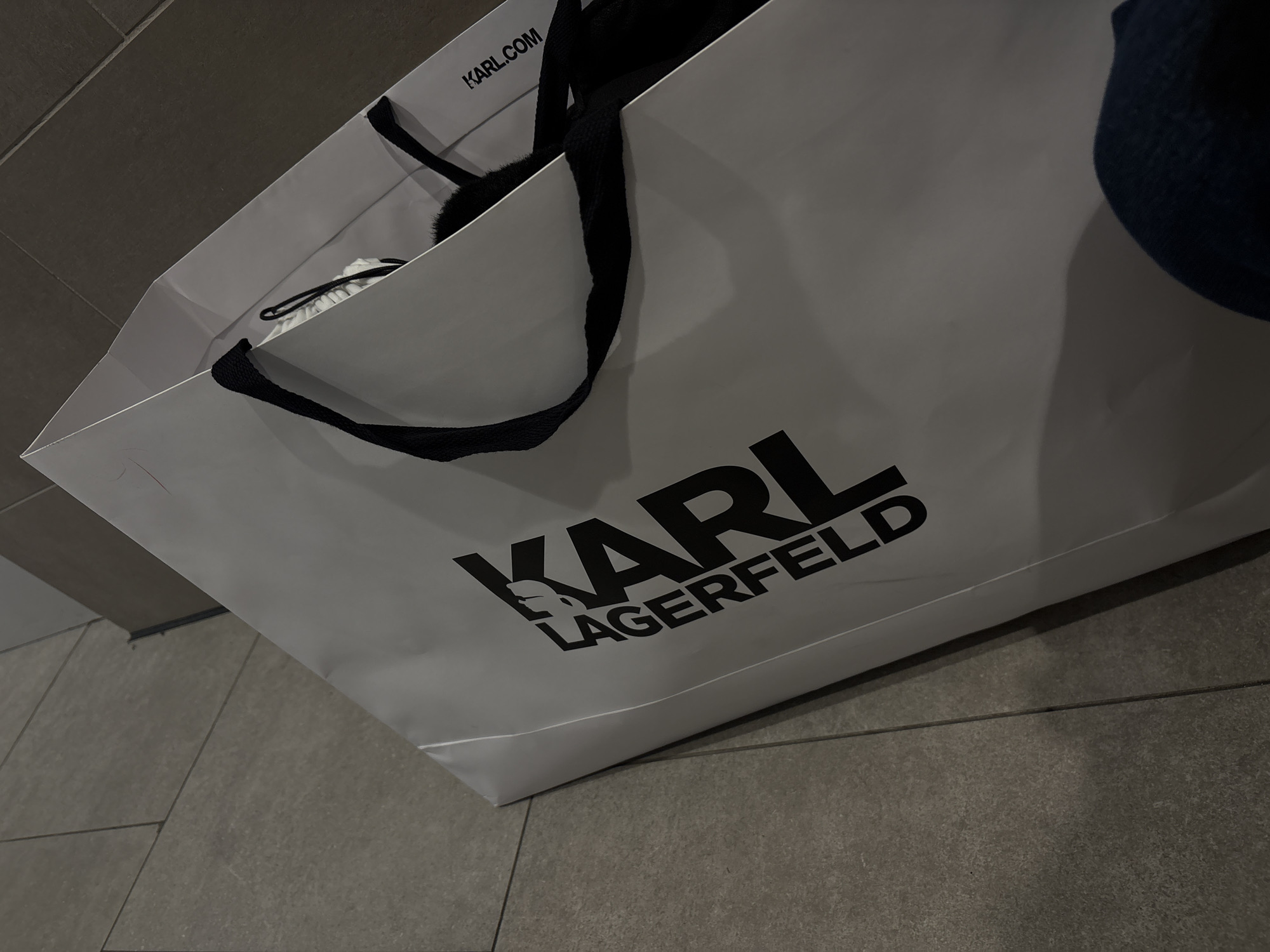 A white Karl Lagerfeld shopping bag sits on a gray tiled floor. The bag has black handles and the brand's logo printed in black. The interior of the bag contains items, including a white object with black drawstrings and a dark-colored item. The background shows a gray wall. No people are visible. There is no definitive information to support the location being Belgrade, Serbia.